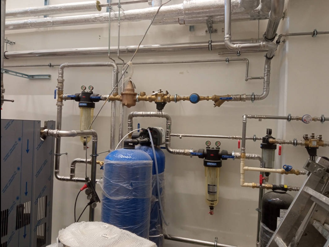 Złożony system rur i filtrów wodnych na ścianie, z niebieskimi zbiornikami i elementami instalacji. Widoczne liczne złącza, zawory i manometry. Część instalacji owinięta folią.