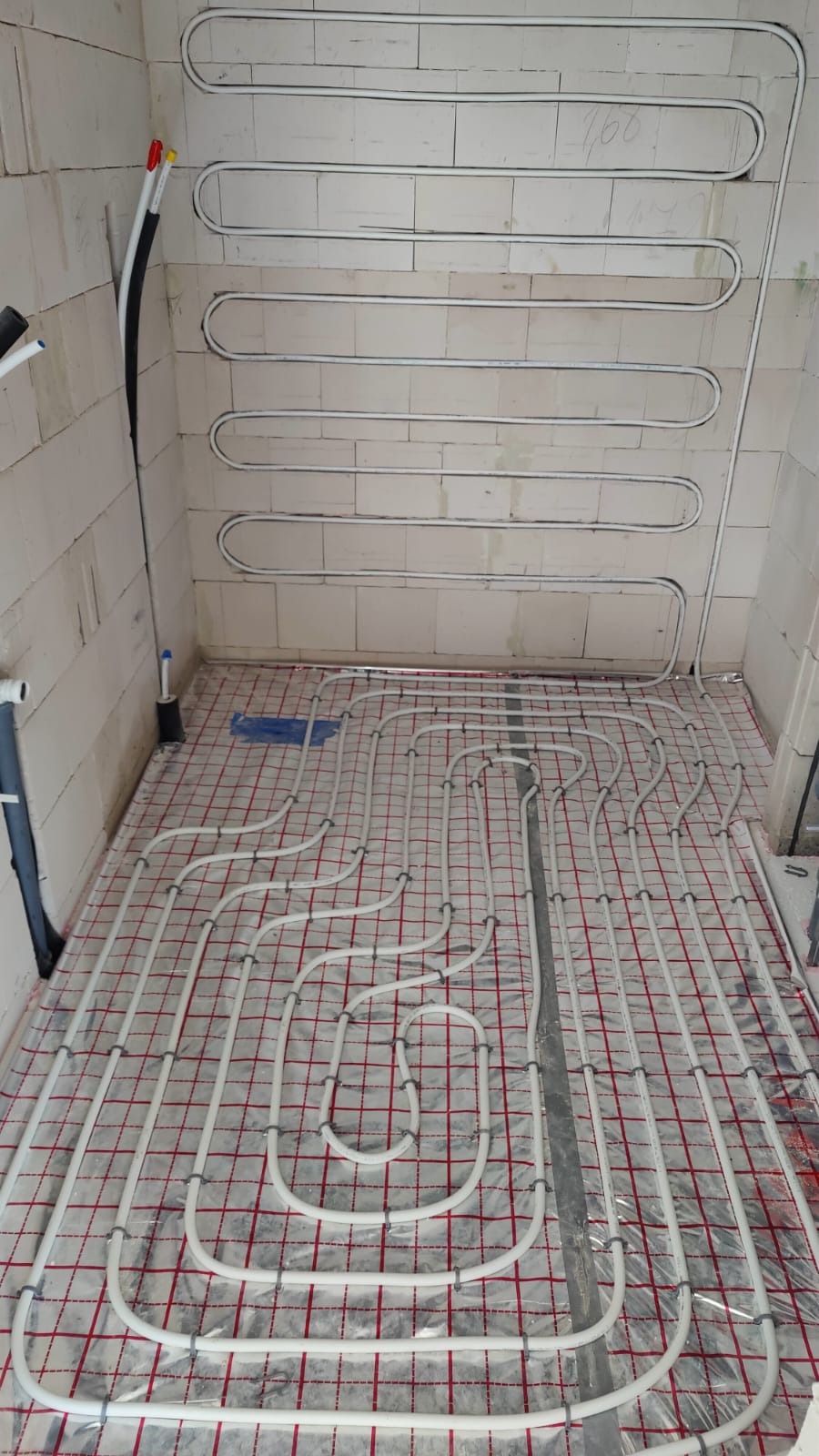 Białe rury ogrzewania podłogowego ułożone na folii z czerwoną kratką w surowym wnętrzu. Rury prowadzone również na ścianie. Widoczne elementy instalacji wodnej.