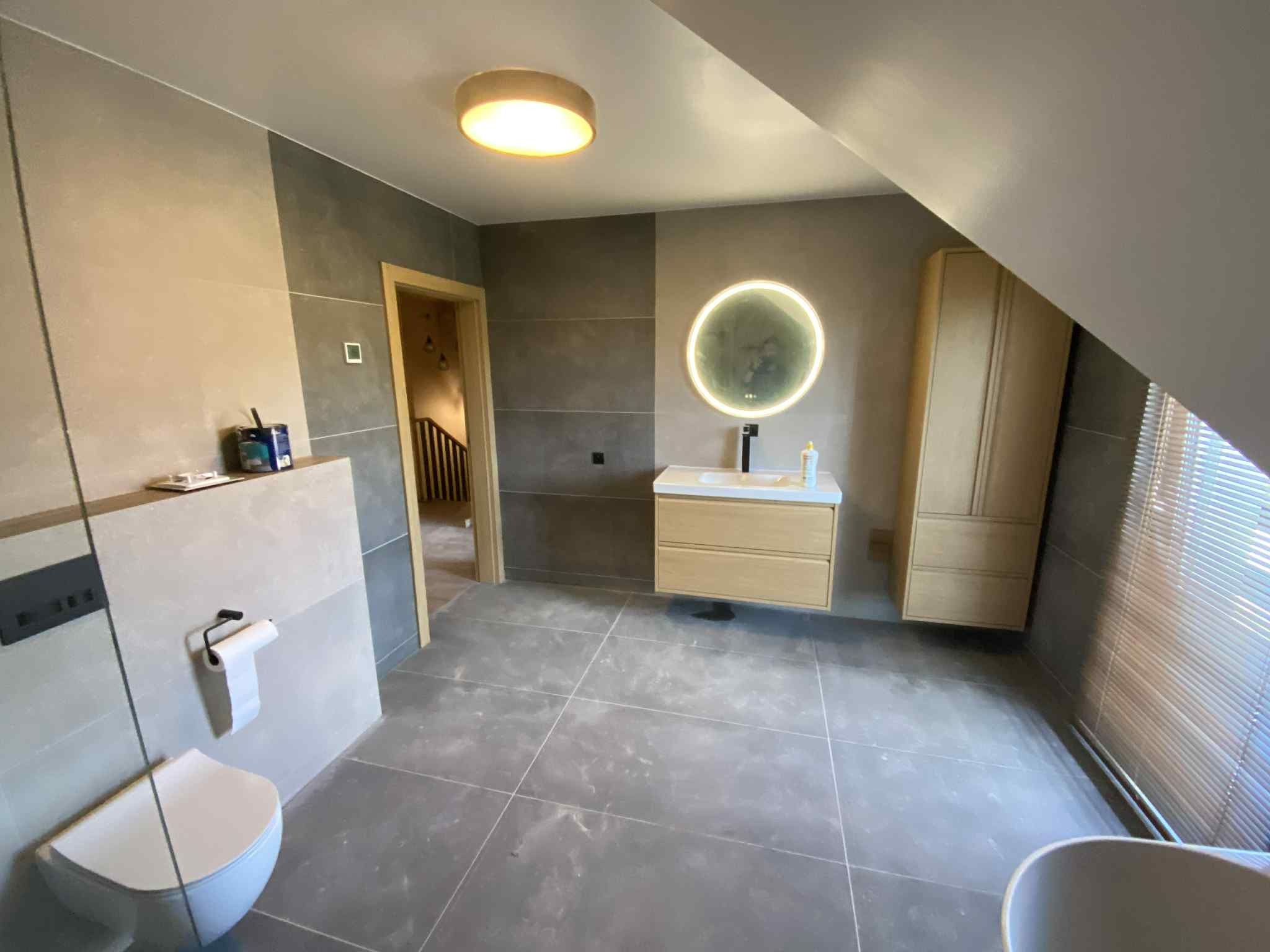 Nowoczesna łazienka w odcieniach szarości z okrągłym lustrem LED i drewnianymi szafkami w Żychlinie. Ukończona aranżacja wnętrza z widoczną armaturą i płytkami.