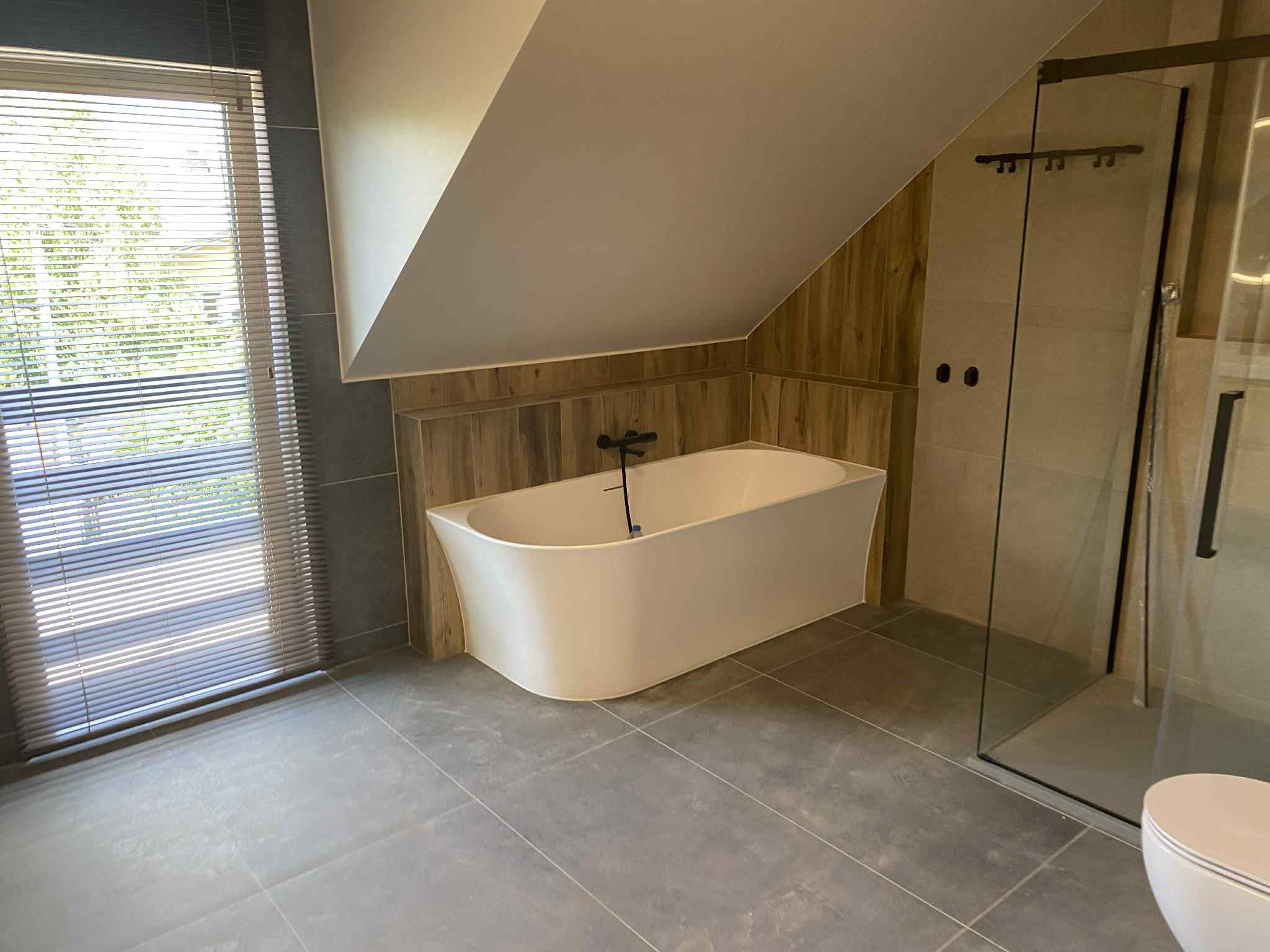 Nowoczesna łazienka na poddaszu z wolnostojącą wanną, kabiną prysznicową ze szklaną ścianą i oknem z żaluzjami. Użyto płytek w odcieniach szarości i drewna.