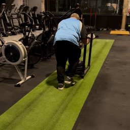 Sick&Flow Krystian Wala - Mężczyzna pcha sanie treningowe po zielonej sztucznej trawie w siłowni. W tle sprzęt do ćwiczeń i okna.