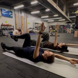 Sick&Flow Krystian Wala - Dwie kobiety ćwiczą na matach w przestronnej siłowni. Wykonują ćwiczenie z uniesionymi rękami, leżąc na plecach. W tle widoczne sprzęty do ćwiczeń i inne osoby.