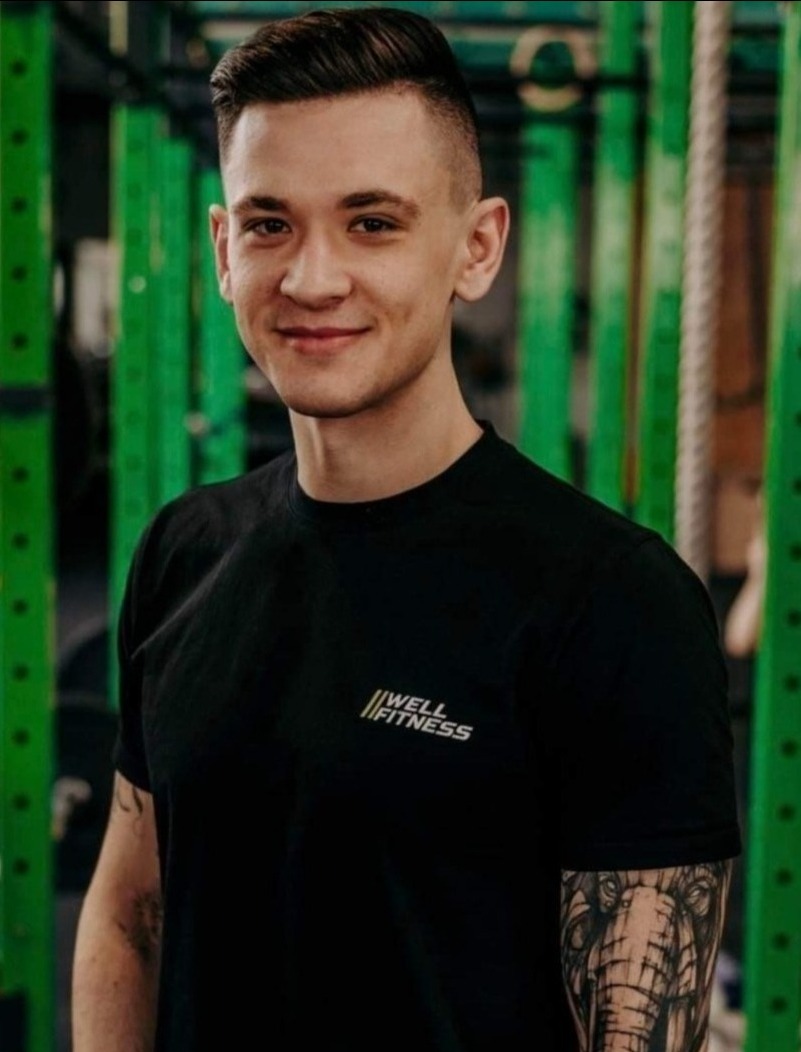 Uśmiechnięty mężczyzna w czarnej koszulce 'Well Fitness' na tle zielonej konstrukcji siłowni. Widoczny tatuaż słonia na ramieniu.