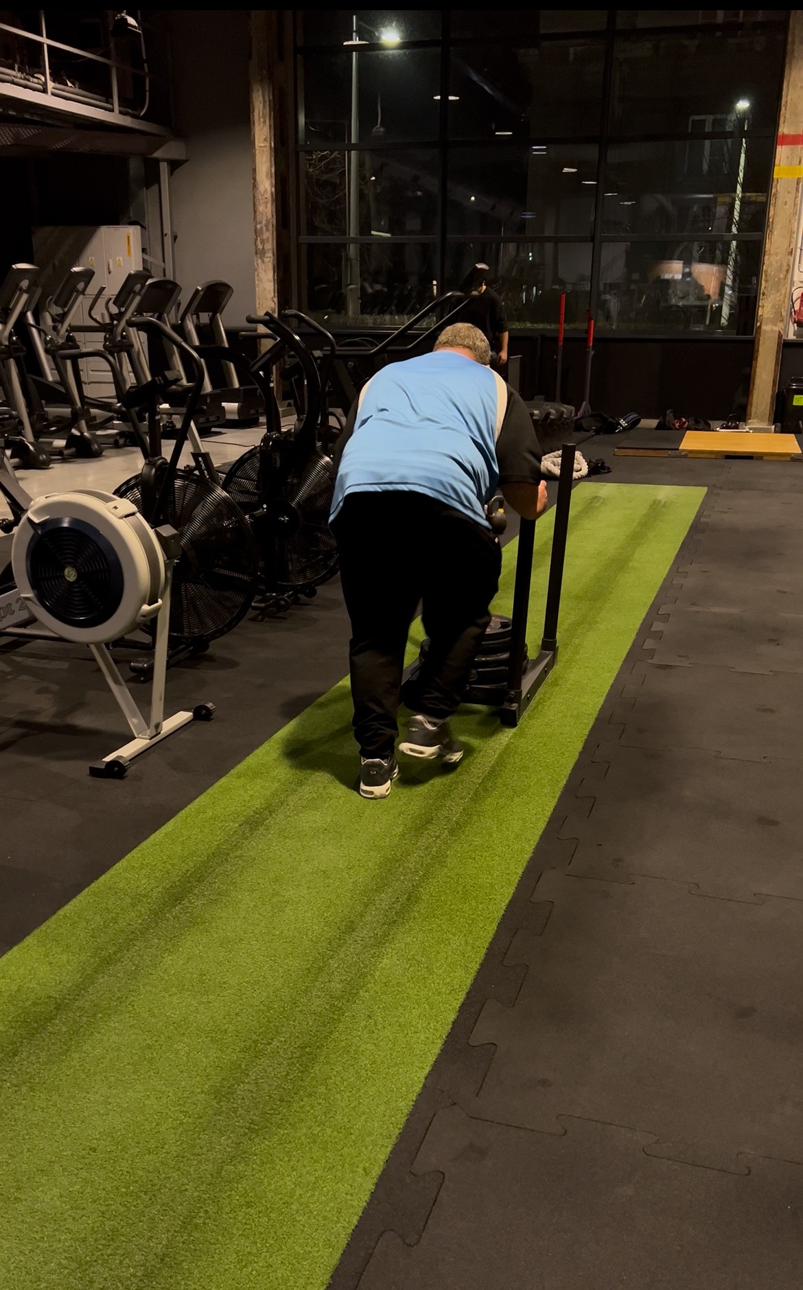 Mężczyzna pcha sanie treningowe po zielonej sztucznej trawie w siłowni. W tle sprzęt do ćwiczeń i okna.