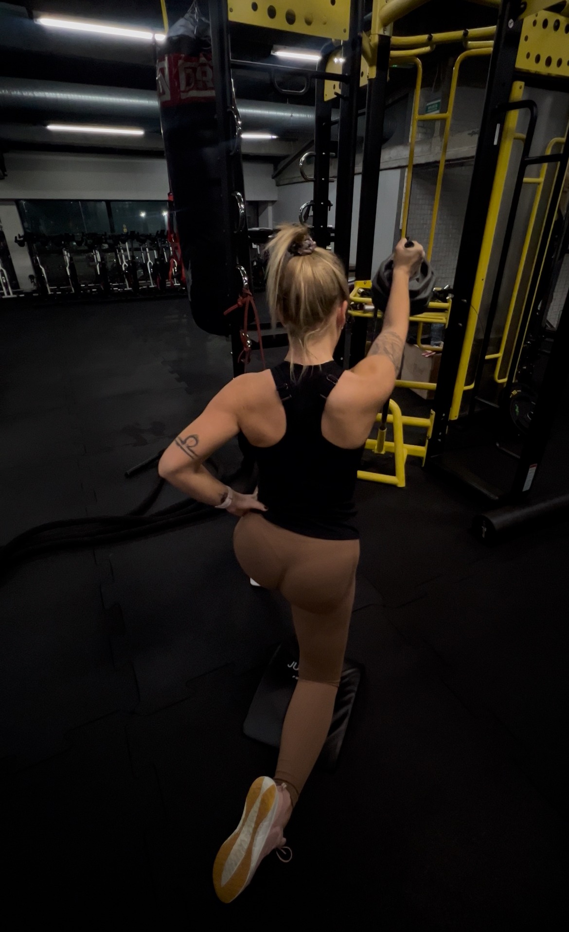 Kobieta w czarnym topie i legginsach wykonuje wykrok z kettlebellem w siłowni. Widoczny worek bokserski i sprzęt do ćwiczeń. Tatuaż na ramieniu.