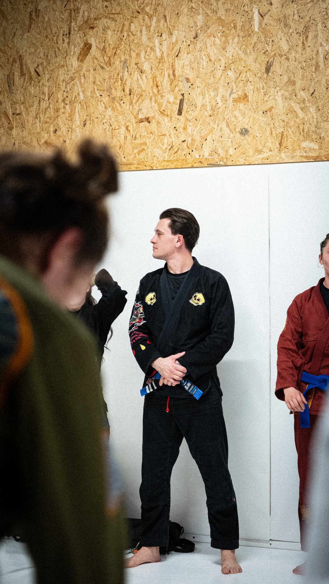 Mężczyzna w czarnym kimonie z niebieskim pasem, stoi na macie do sportów walki, z boku widoczni inni uczestnicy treningu. W tle ściana z płyty OSB.