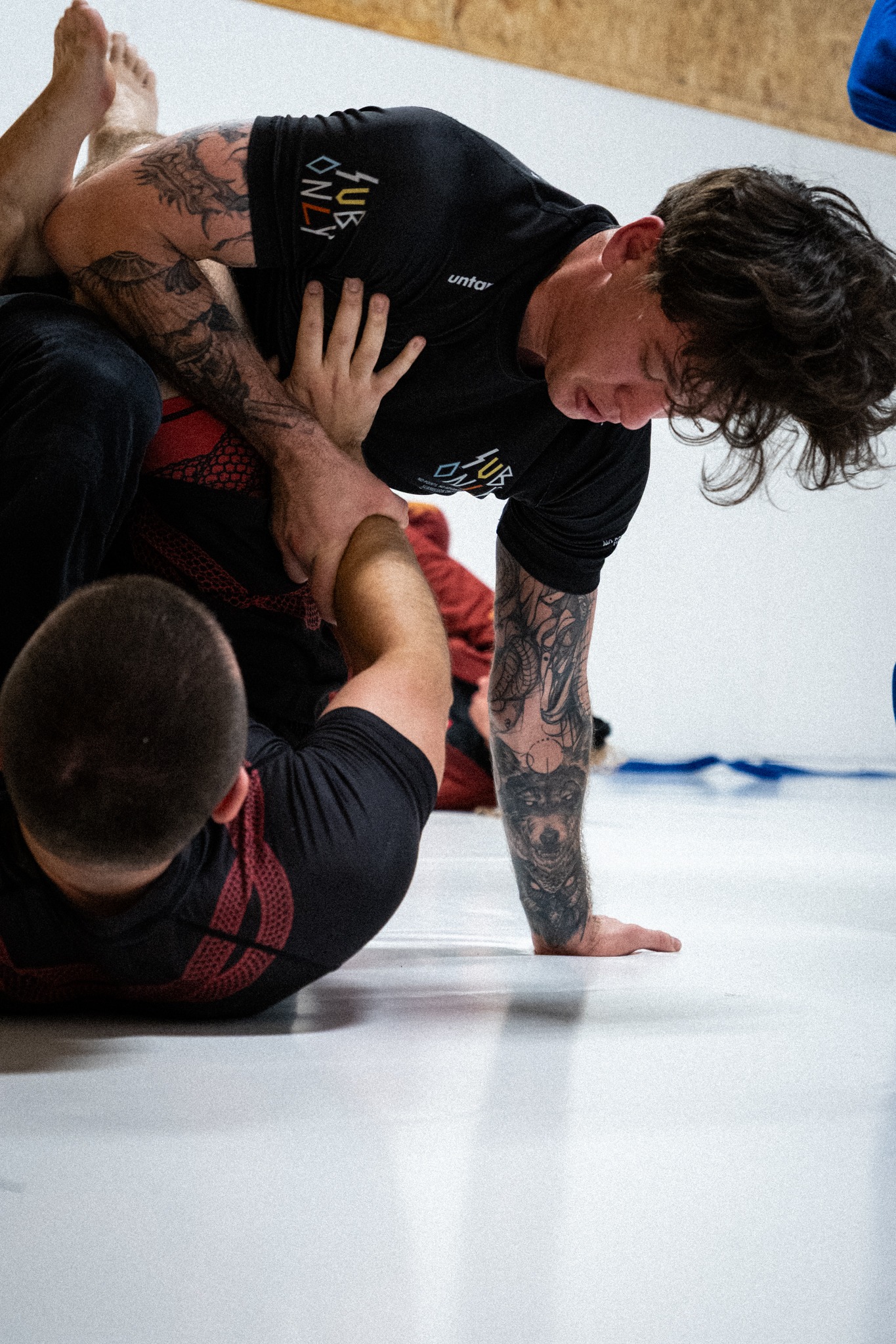 Trening BJJ w Bielsko-Białej: Dwóch mężczyzn w trakcie walki na macie. Jeden z tatuażami na rękach, w czarnej koszulce, dominuje nad przeciwnikiem w czerwono-czarnej koszulce.