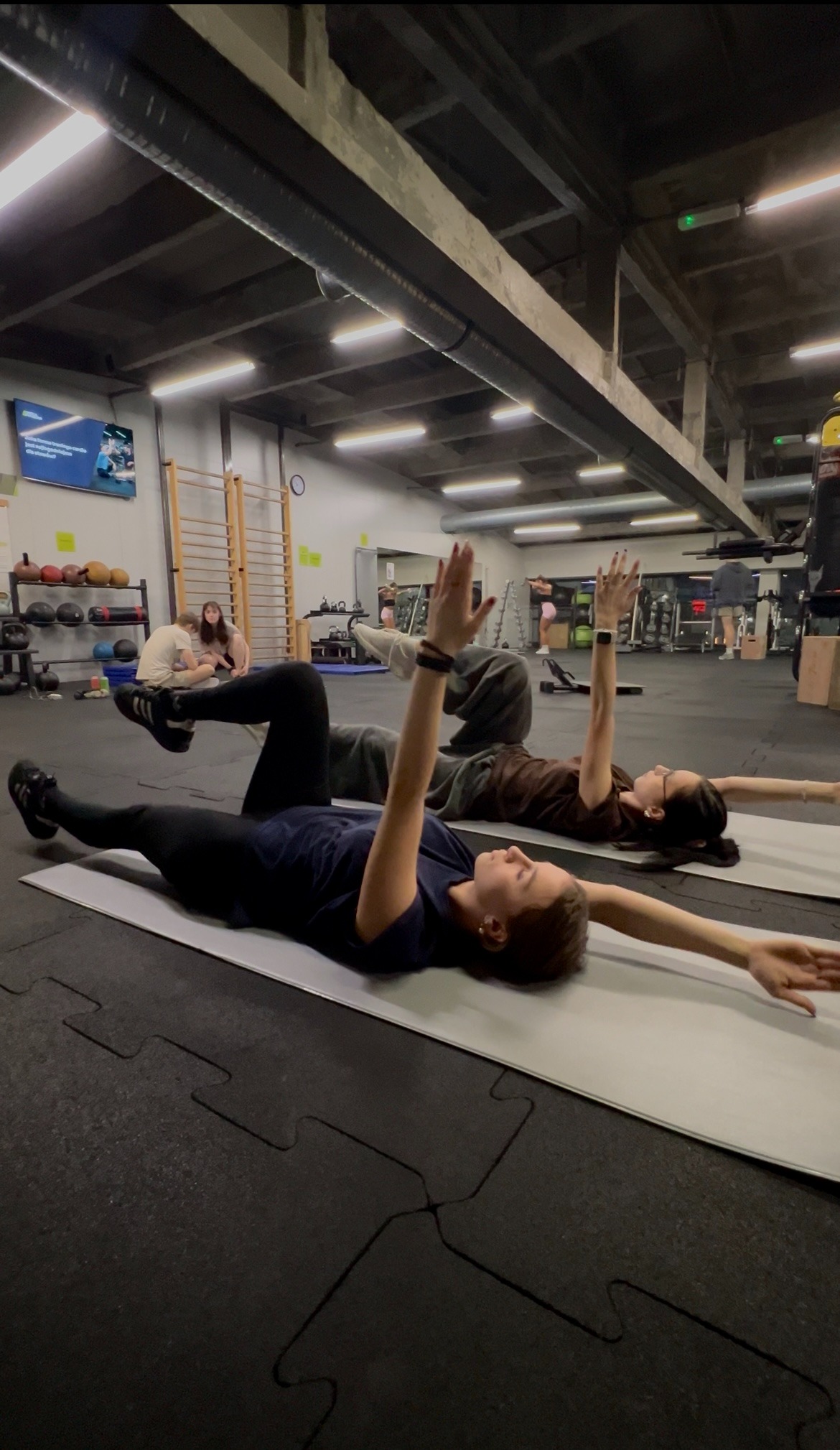 Dwie kobiety ćwiczą na matach w przestronnej siłowni. Wykonują ćwiczenie z uniesionymi rękami, leżąc na plecach. W tle widoczne sprzęty do ćwiczeń i inne osoby.