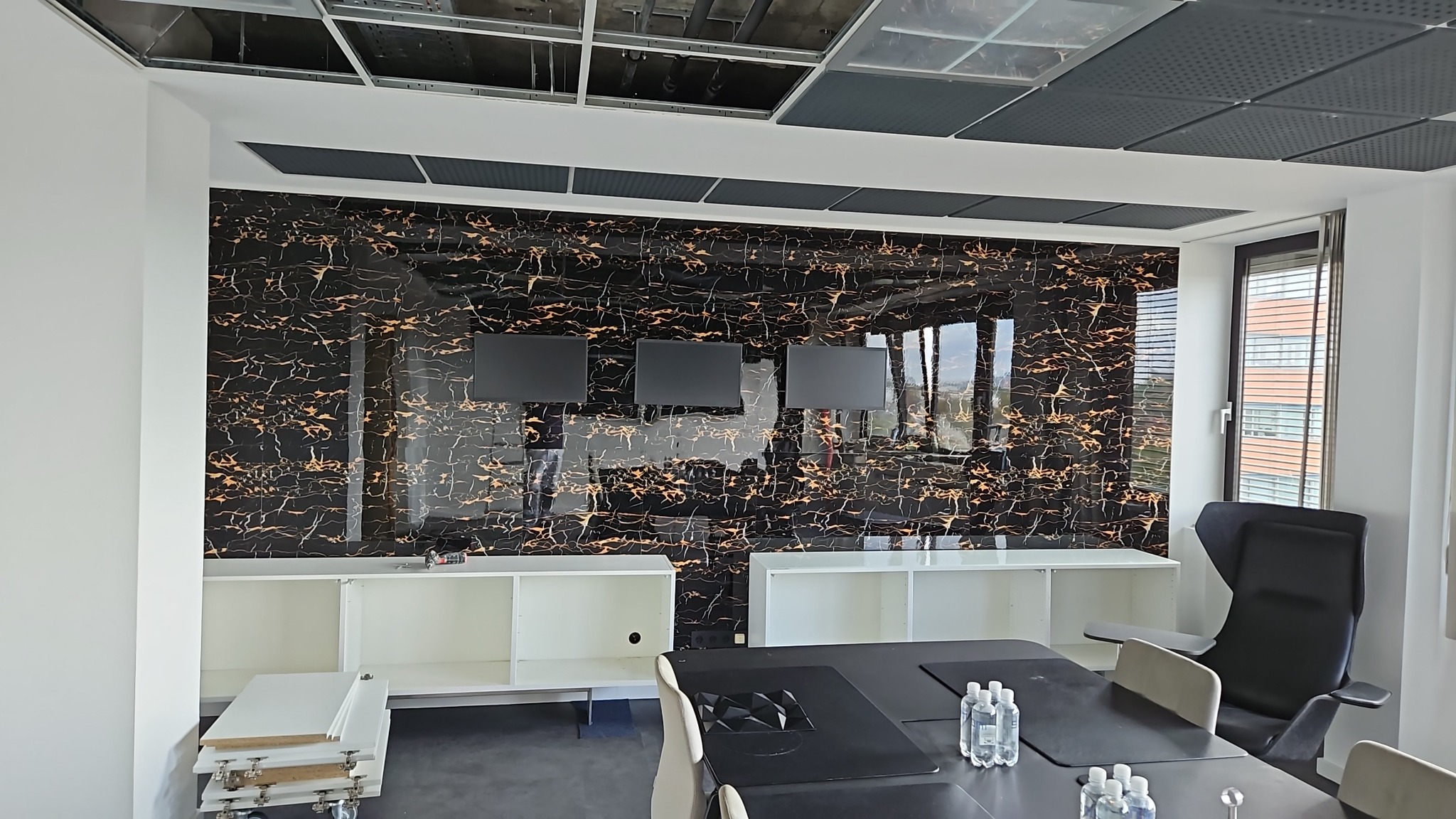 Elegancka sala konferencyjna z czarną ścianą akcentową w marmurkowy wzór, trzema monitorami i białymi szafkami. Widoczny fragment sufitu podwieszanego i stół konferencyjny.