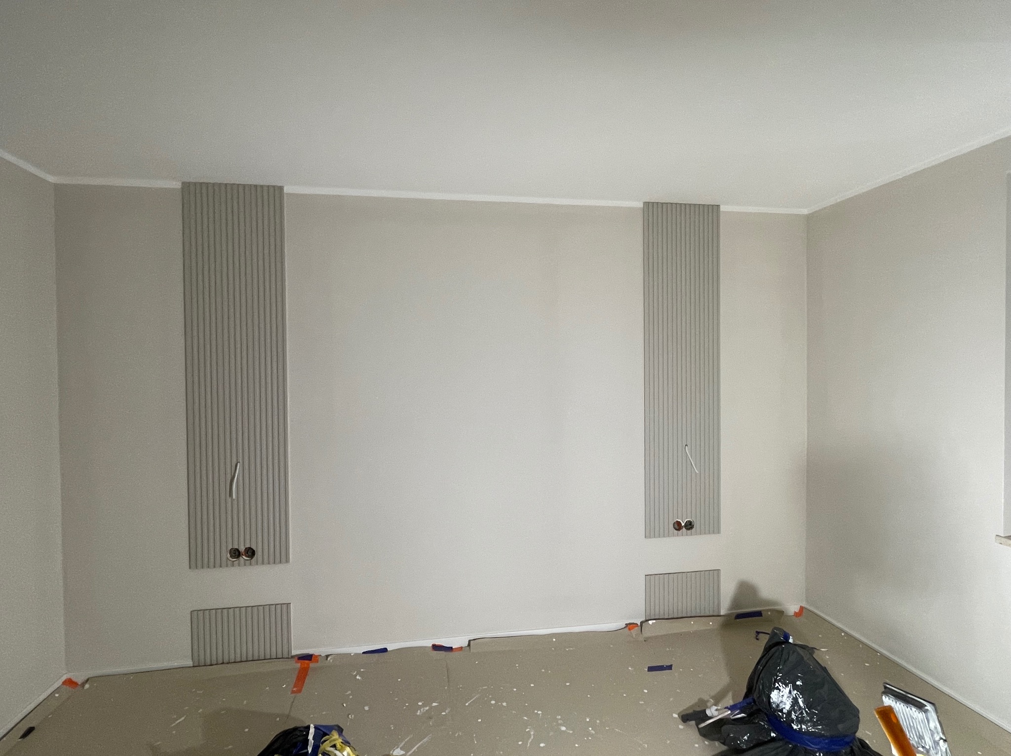 Jasne wnętrze w trakcie wykańczania: pionowe panele dekoracyjne na ścianie, zabezpieczona podłoga, widoczne gniazdka elektryczne i kable. Minimalistyczny design w neutralnej kolorystyce.