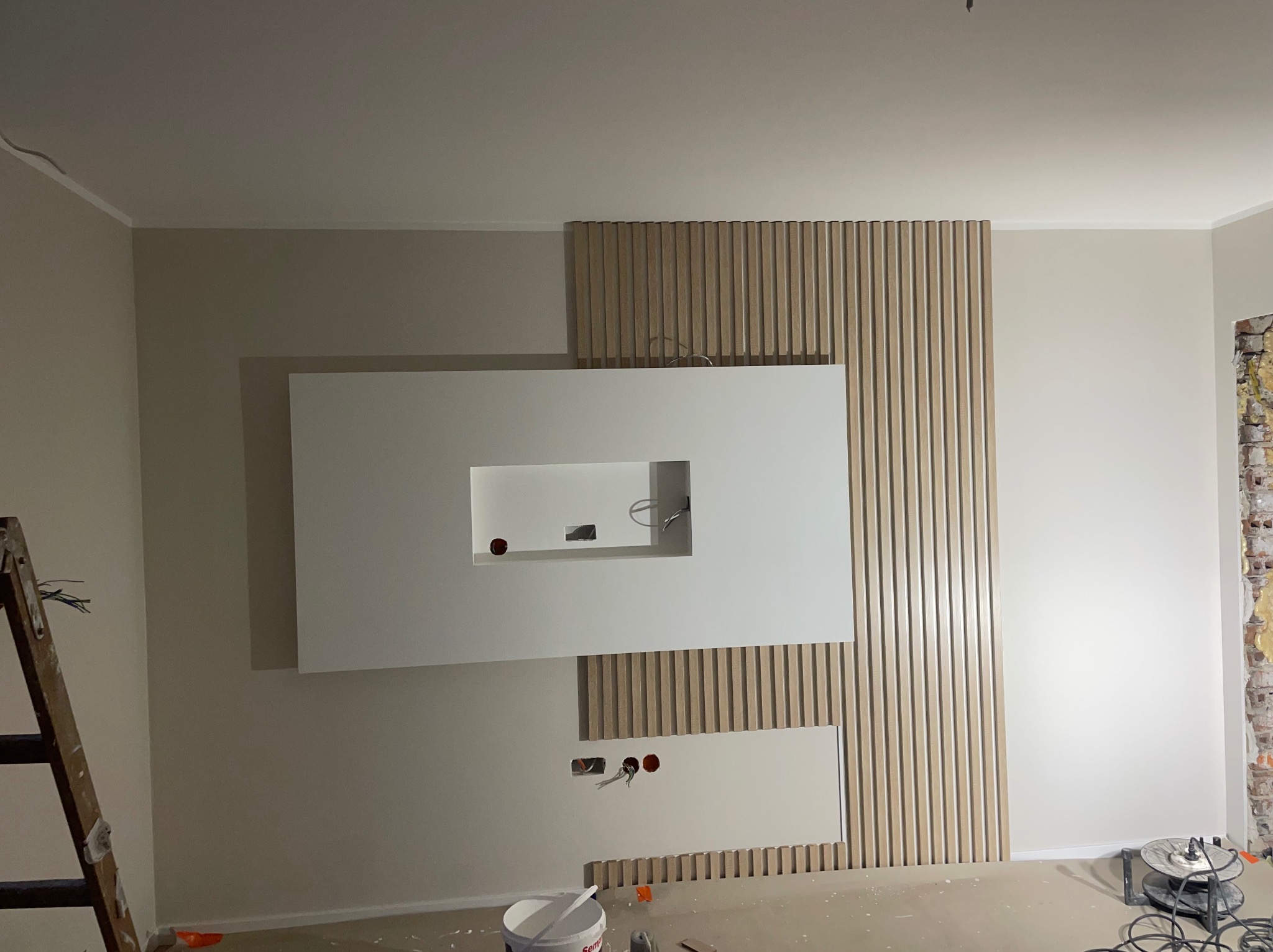 Ściana z drewnianymi lamelami i białą zabudową pod TV w trakcie montażu. Widoczne kable i puszki elektryczne. Drabina oparta o ścianę. Podłoga zabezpieczona folią.