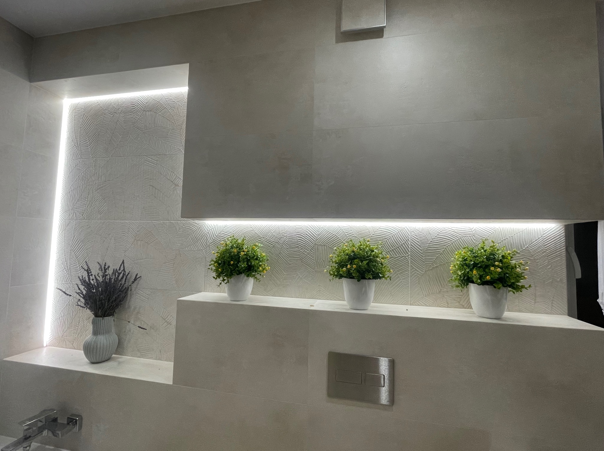 Nowoczesna łazienka z szarymi płytkami, podświetleniem LED i dekoracyjnymi roślinami w doniczkach. Minimalistyczny design z akcentami natury i subtelnym oświetleniem.