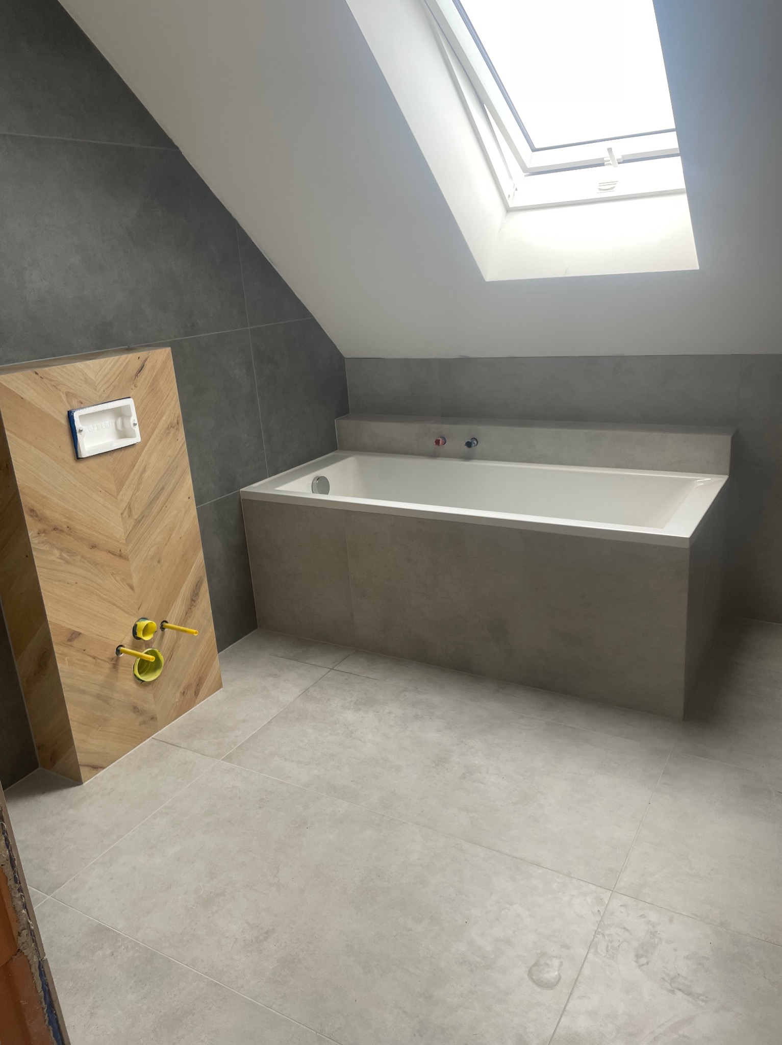 Nowoczesna łazienka na poddaszu z oknem dachowym. Ukończona zabudowa wanny w szarym odcieniu, drewniana zabudowa stelaża WC z wzorem jodełki. Szare płytki na ścianach i podłodze.