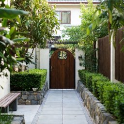 MILA SPÓŁKA Z OGRANICZONĄ ODPOWIEDZIALNOŚCIĄ - Ujęcie na ścieżkę z płyt prowadzącą do drewnianej furtki z ozdobnym okienkiem, otoczonej równo przyciętymi żywopłotami i kamiennymi murkami w zadbanym ogrodzie.