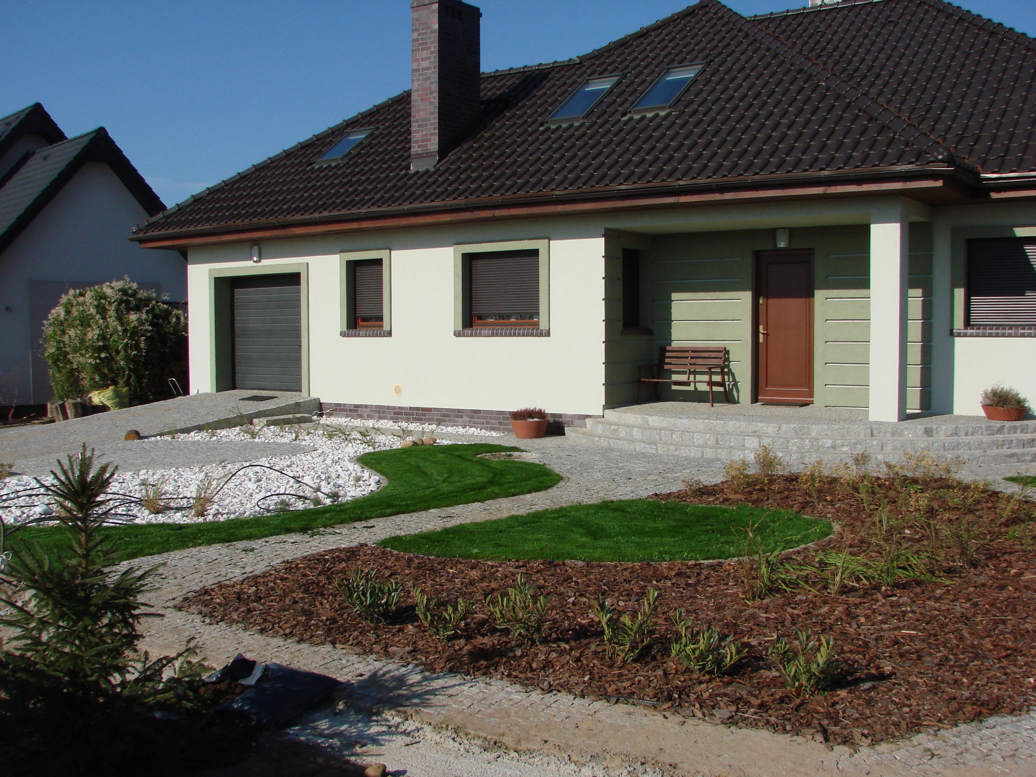 Nowoczesny dom z zadbanym ogrodem: trawnik, białe kamienie, kora. Ścieżka z kostki brukowej prowadzi do wejścia. Widoczne elementy systemu nawadniania.