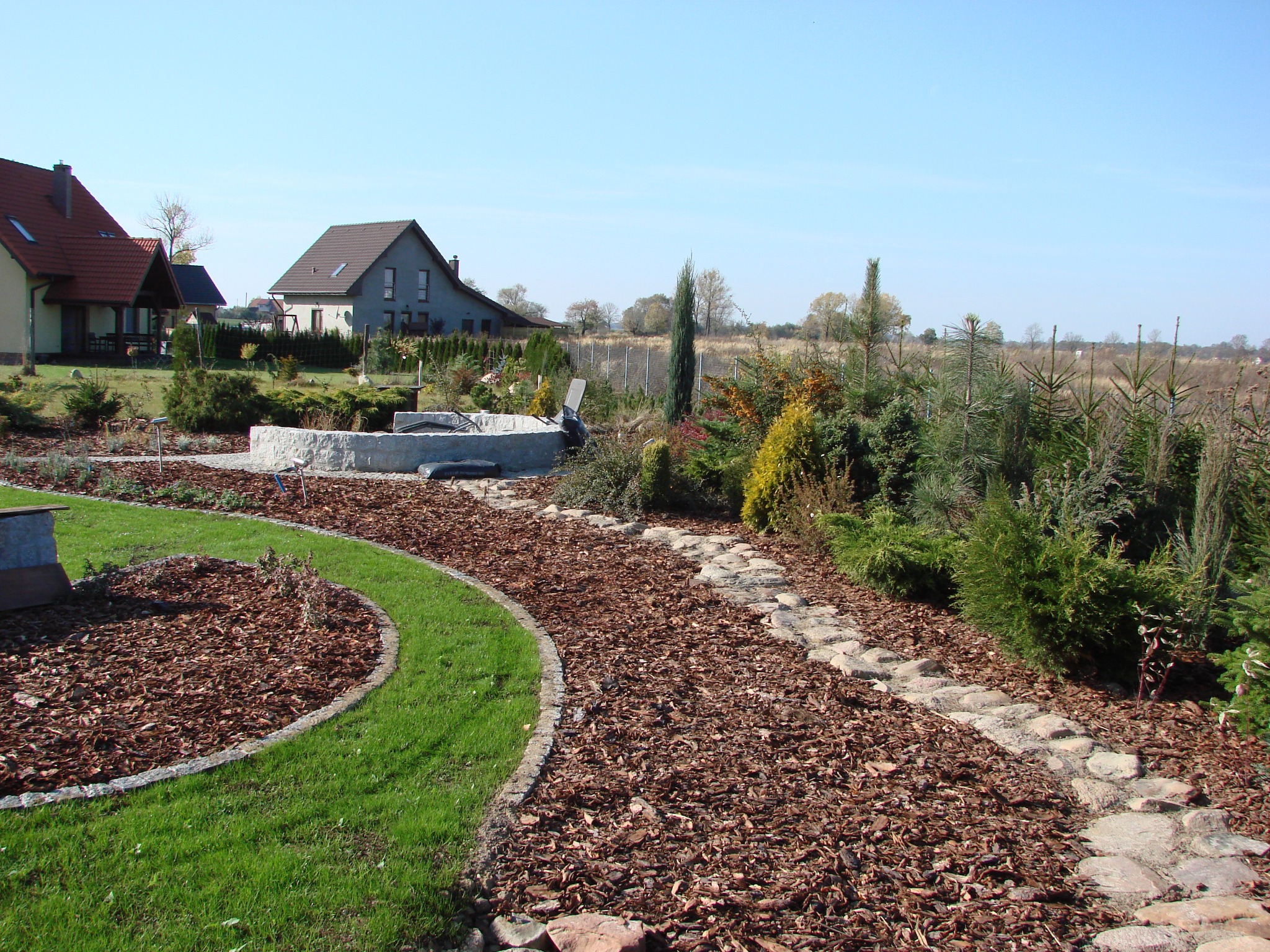 Ogród z ścieżką z kamieni i kory, obrzeża trawnikowe z kostki, w tle domy i drzewa iglaste. Słoneczny dzień, błękitne niebo. Widok na zadbany ogród.