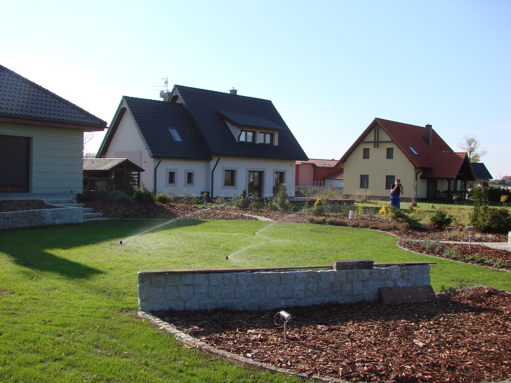 Zadbany trawnik z systemem nawadniania przed domem. Widoczne zraszacze w akcji, rabaty z korą i kamienny murek. W tle zabudowa jednorodzinna w słoneczny dzień.