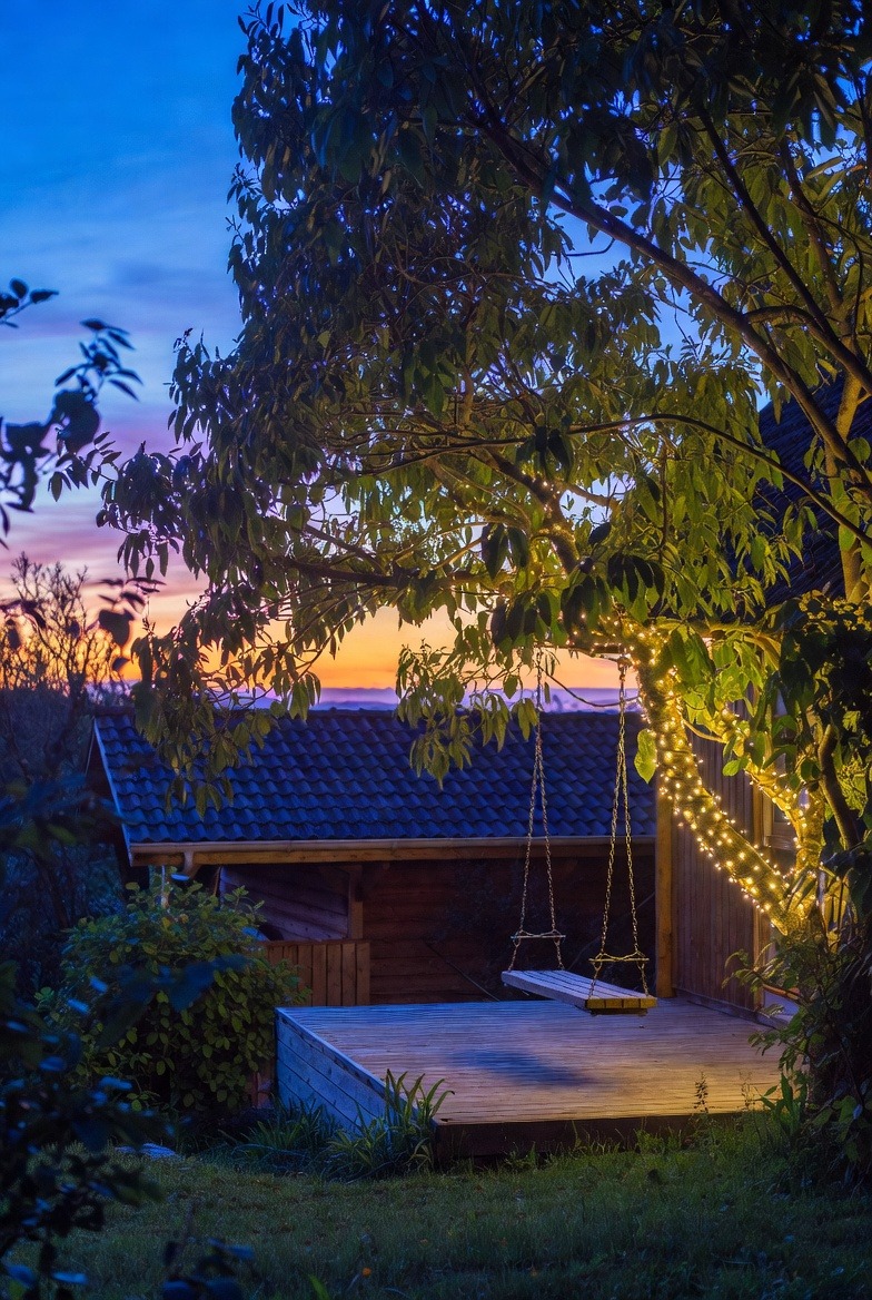 Oświetlony ogród z huśtawką na tle zachodzącego słońca. Drewniany taras i budynek w tle. Drzewo oplecione lampkami tworzy przytulny klimat.
