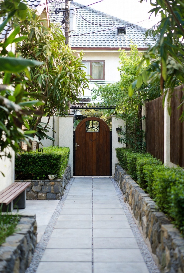 Ujęcie na ścieżkę z płyt prowadzącą do drewnianej furtki z ozdobnym okienkiem, otoczonej równo przyciętymi żywopłotami i kamiennymi murkami w zadbanym ogrodzie.