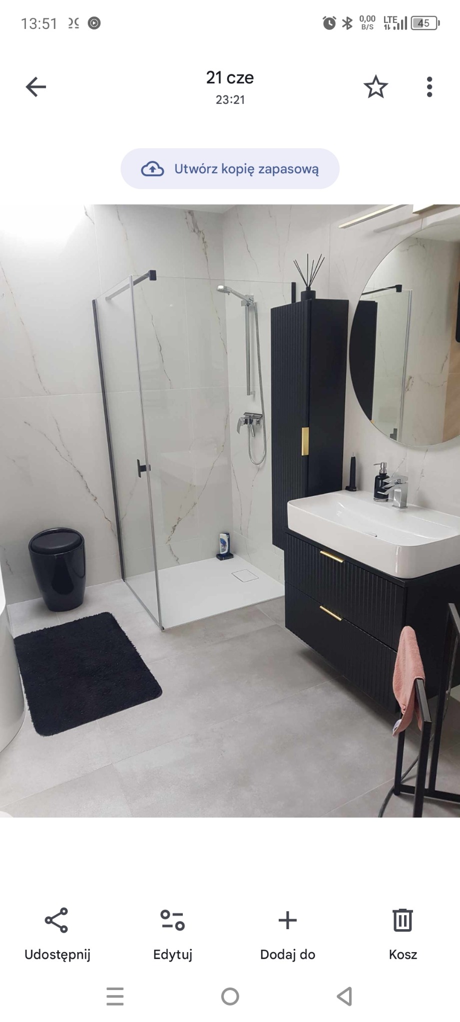 Nowoczesna łazienka w Zielonce: kabina prysznicowa typu walk-in z czarnymi detalami, białe płytki z marmurowym wzorem, czarna szafka z umywalką i okrągłe lustro.