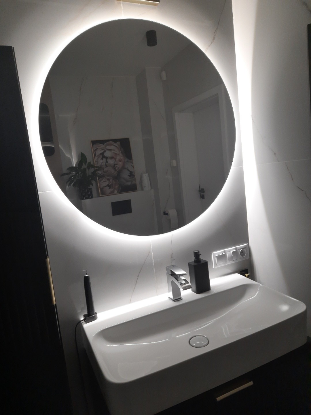 Nowoczesna łazienka: biała umywalka z czarną szafką, okrągłe lustro z podświetleniem LED, marmurowe płytki. Minimalistyczny design, eleganckie wykończenie.