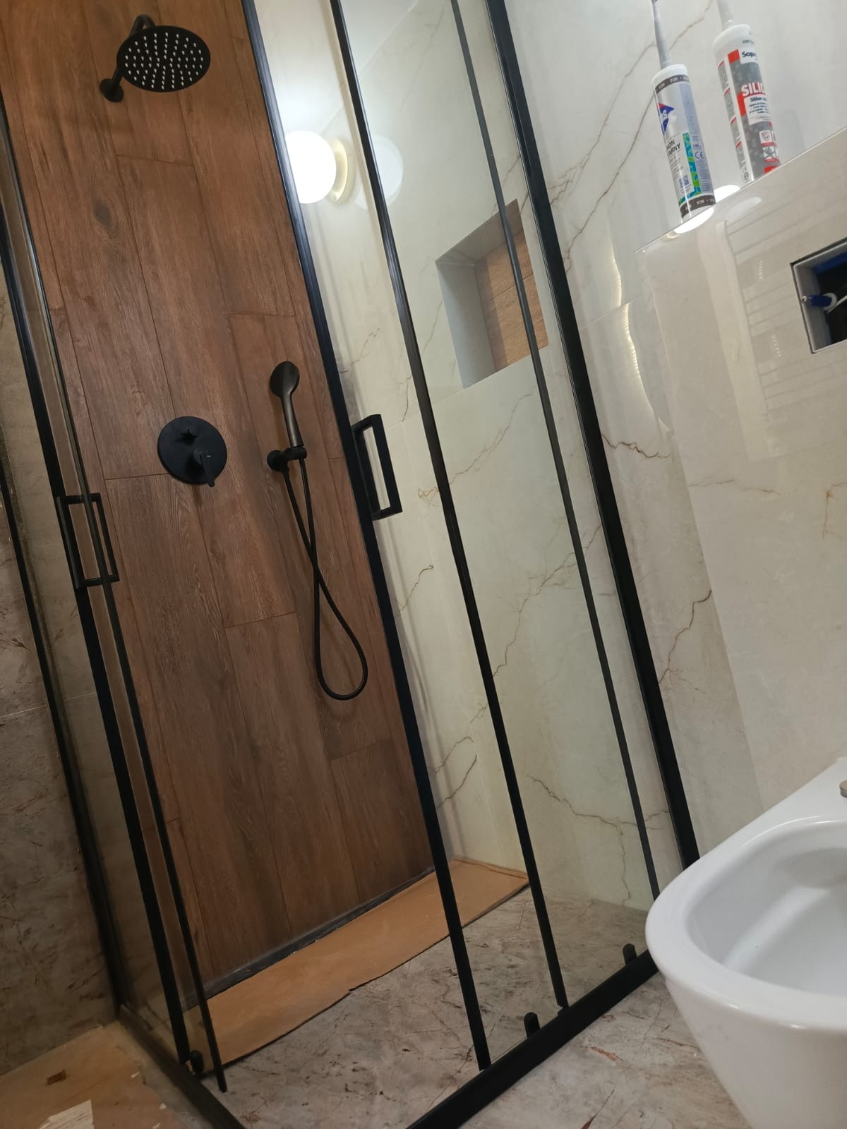Nowoczesna kabina prysznicowa z drewnopodobnymi panelami i czarną armaturą, marmurowe płytki na ścianach, minimalistyczny design łazienki.