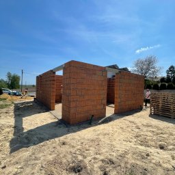 BUD-KAM Uslugi budowlane Krystian Kamyszek - Wznoszone ściany z czerwonej cegły dziurawki na fundamencie, widoczne zbrojenia, palety i pracownik w tle, w słoneczny dzień na placu budowy.