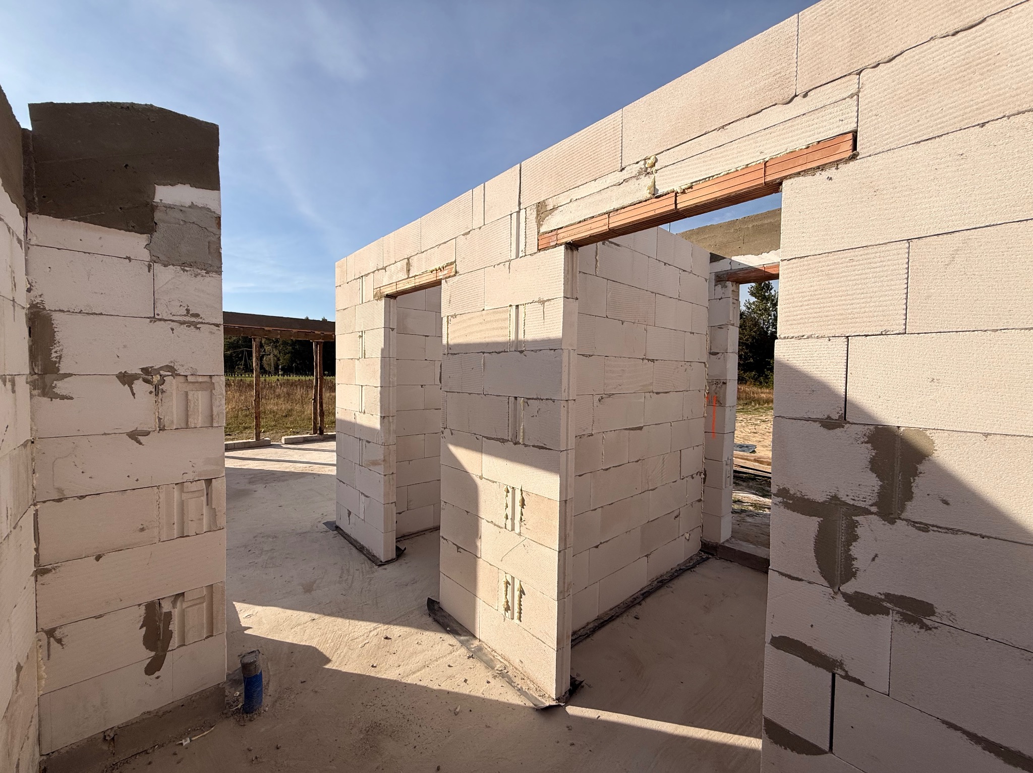 Wnętrze domu w trakcie budowy: ściany z białych bloczków, widoczne otwory drzwiowe i okienne, świeży beton, w tle błękitne niebo. Budowa domu w stanie surowym otwartym.