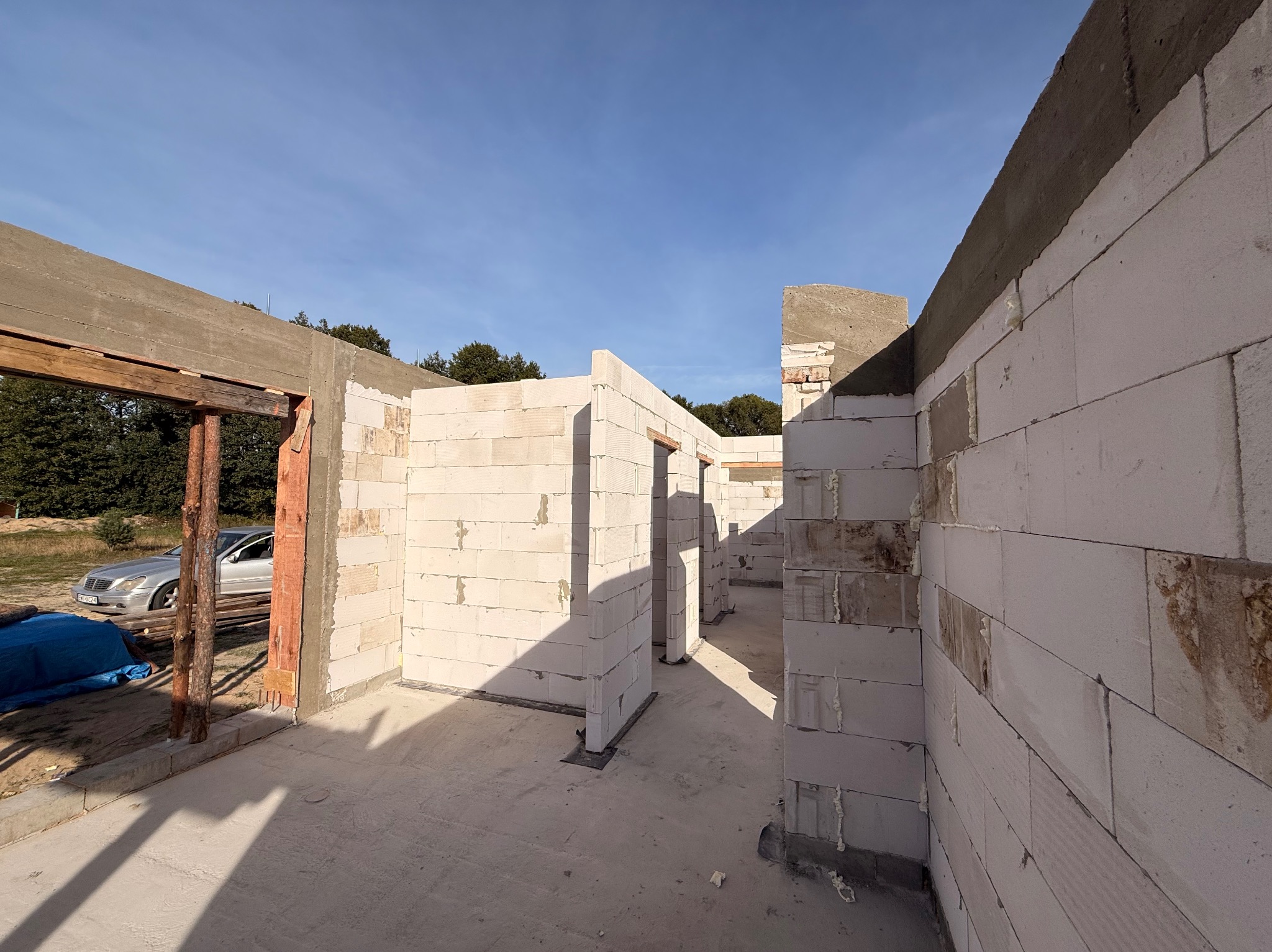 Wnętrze domu w trakcie budowy: ściany z białych bloczków, widoczne otwory drzwiowe i okienne, częściowo zabetonowany wieniec. Na zewnątrz zaparkowany srebrny samochód.