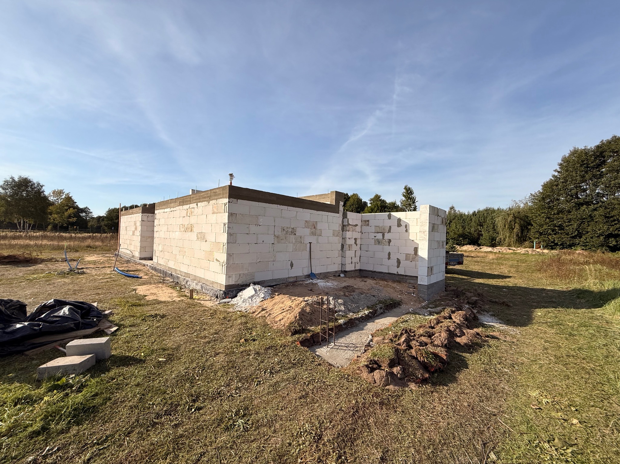 Wznoszenie ścian parterowego domu z białych bloczków, widoczne zbrojenie fundamentów. Budowa w toku, na zewnątrz trawa i drzewa, błękitne niebo.
