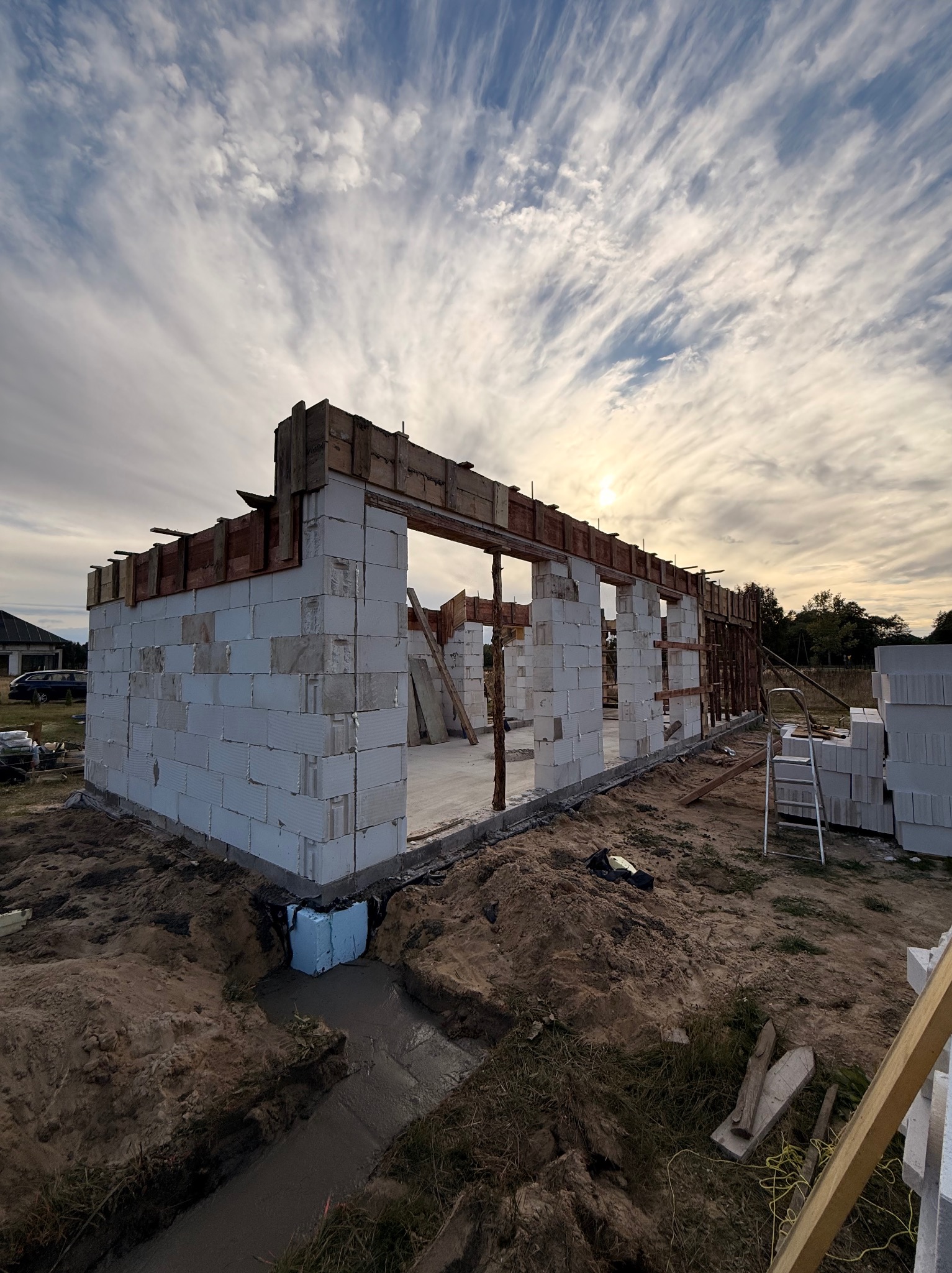 Wznoszenie ścian domu z bloczków betonowych, widoczne szalunki i fundamenty. Na pierwszym planie wykop z betonem. W tle błękitne niebo z chmurami.