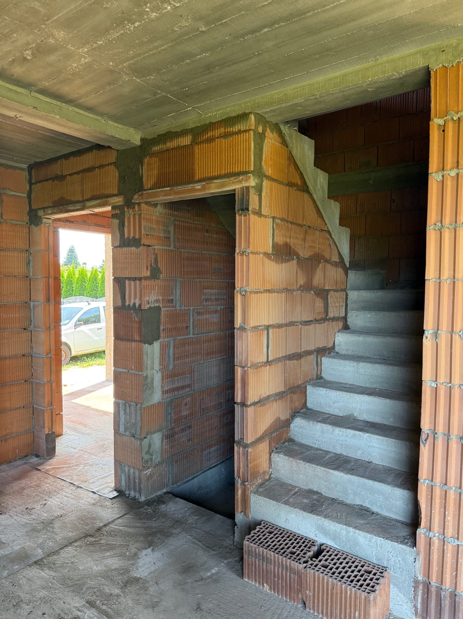 Wnętrze w trakcie budowy: ściany z czerwonej cegły, betonowe schody prowadzące na górę, surowy betonowy strop, widok na zewnątrz przez otwór drzwiowy.