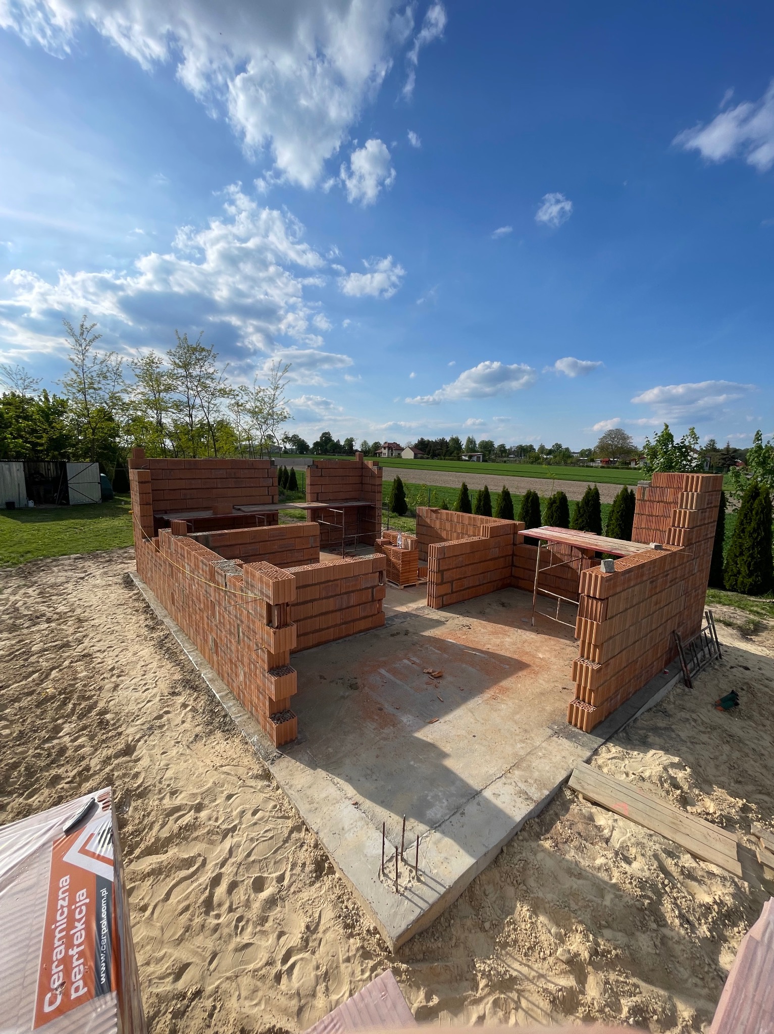 Wznoszenie ścian z czerwonej cegły na fundamencie, widok z góry. Częściowo zbudowana konstrukcja na tle błękitnego nieba z chmurami i zieleni. Widoczne rusztowania i paleta cegieł.