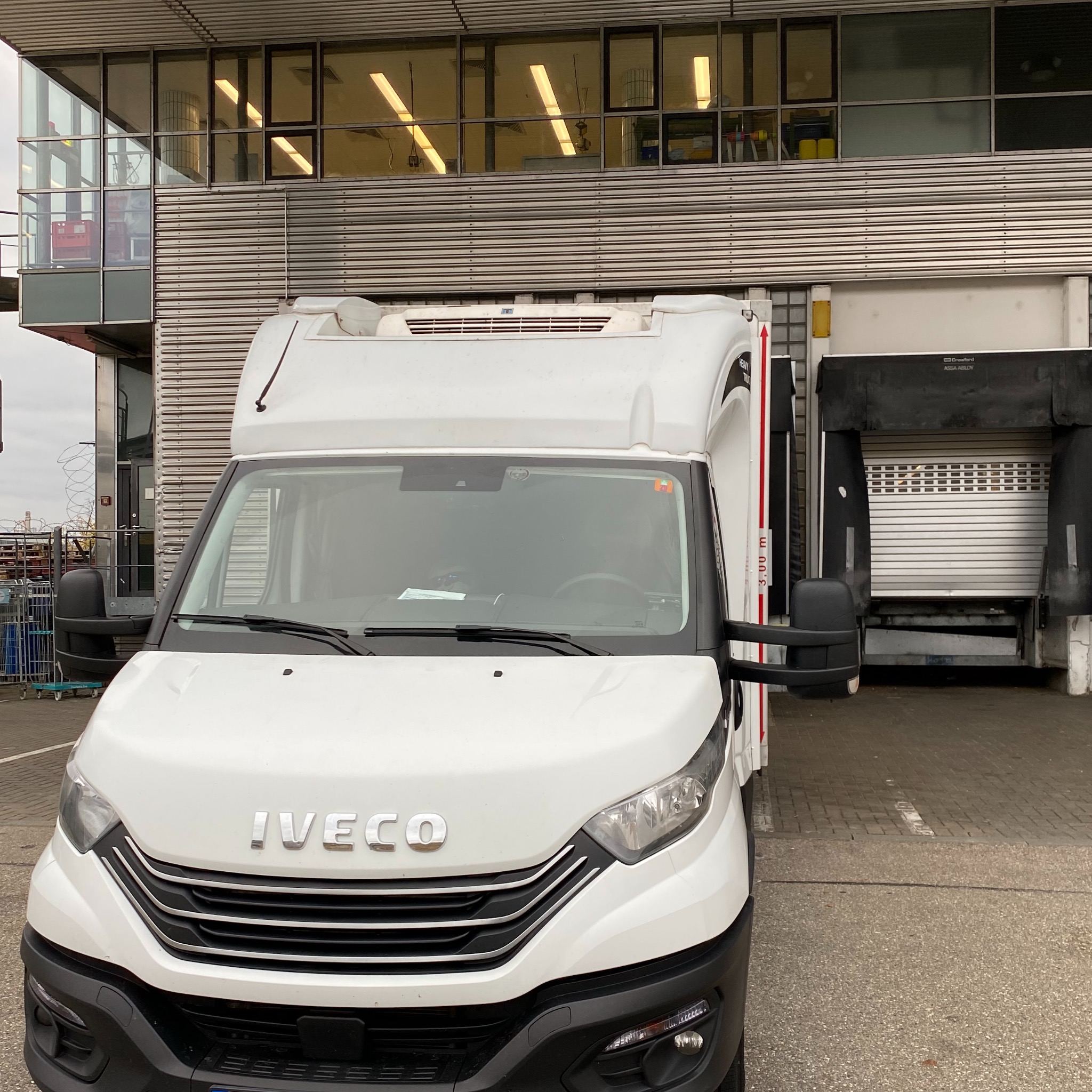 Biały samochód dostawczy Iveco zaparkowany przed budynkiem z rampą załadunkową. Widoczne detale pojazdu i otoczenie przemysłowe.