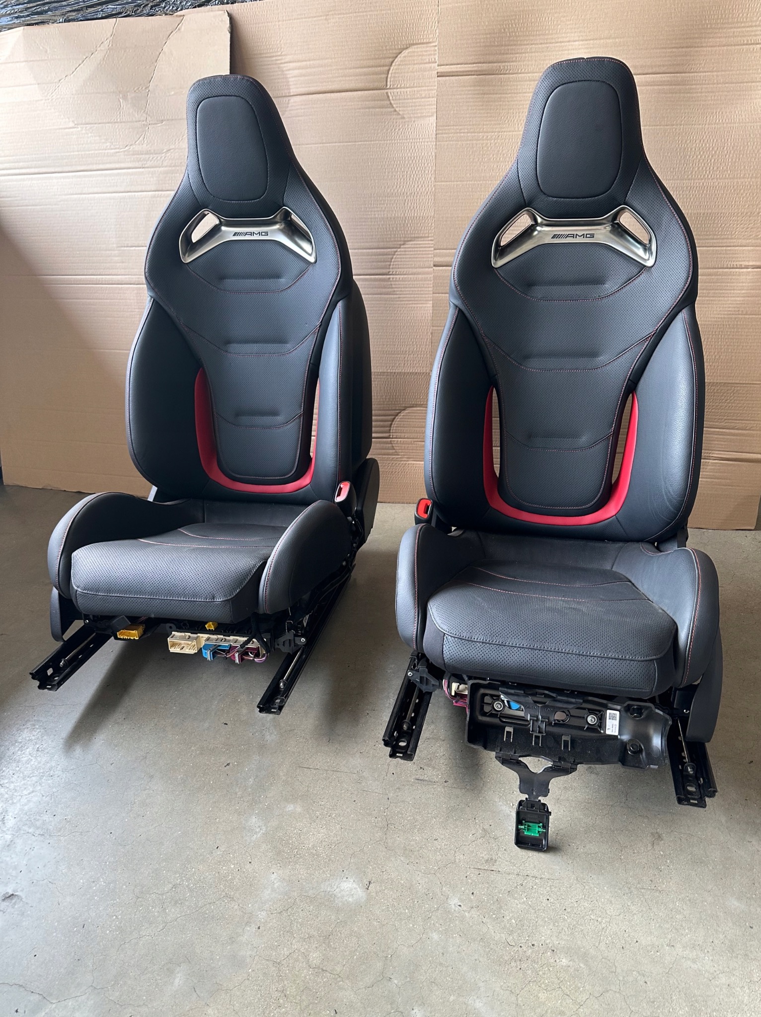 Dwa czarne fotele samochodowe AMG z czerwonymi akcentami, widoczne od przodu na szarym tle. Fotele z widocznymi elementami montażowymi i przewodami.