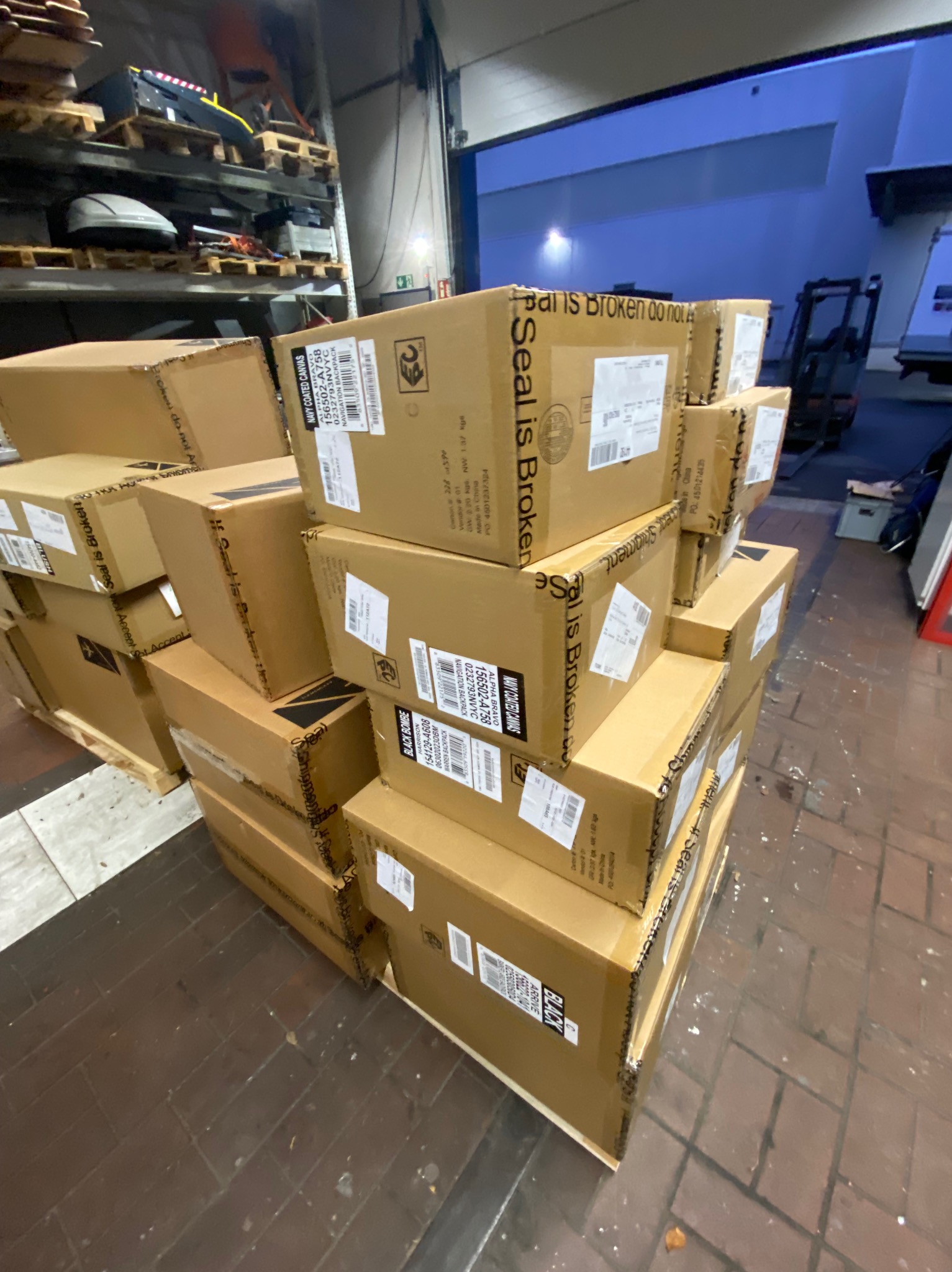 Stos kartonowych pudeł z etykietami w magazynie, gotowych do transportu. Widoczne regały z towarem w tle. Usługa transportowa.