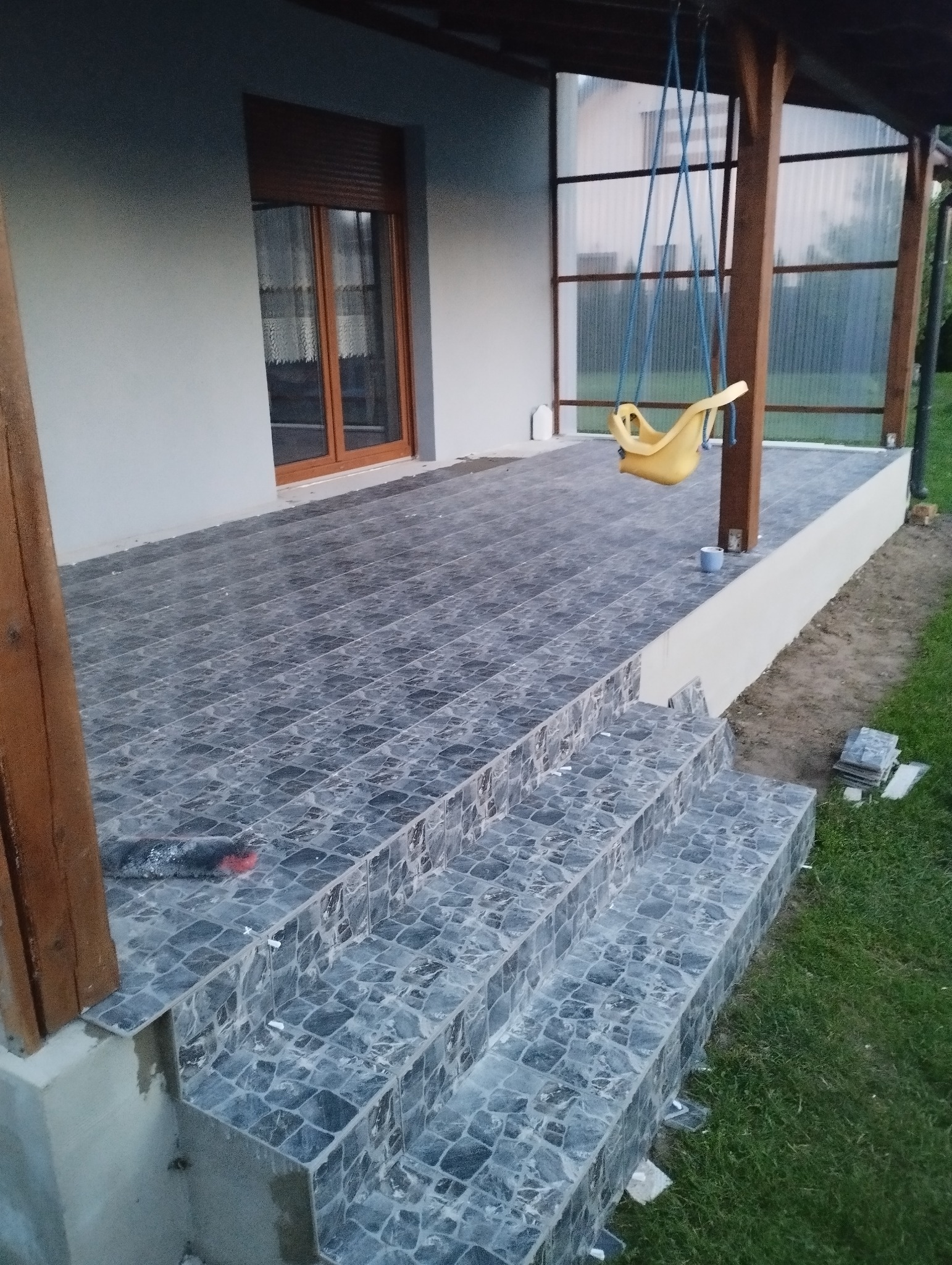 Wykończony taras z szarych płytek mozaikowych, schody z płytek, huśtawka dla dziecka, drewniane słupy podtrzymujące zadaszenie, okno z roletą zewnętrzną.