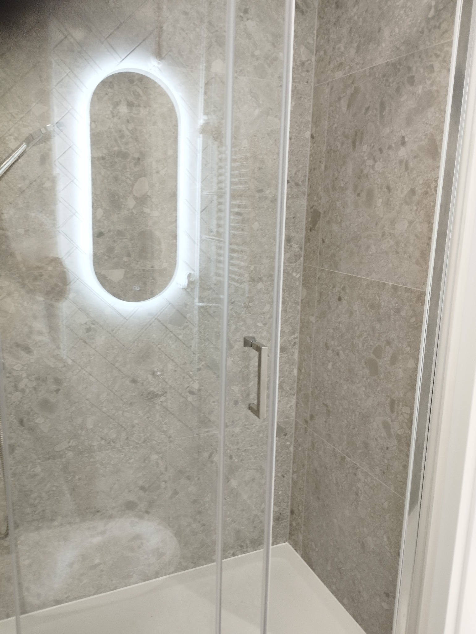 Szklana kabina prysznicowa z podświetlanym lustrem w kształcie łuku na szarej, wzorzystej ścianie. Widoczny fragment baterii prysznicowej i uchwyt kabiny.