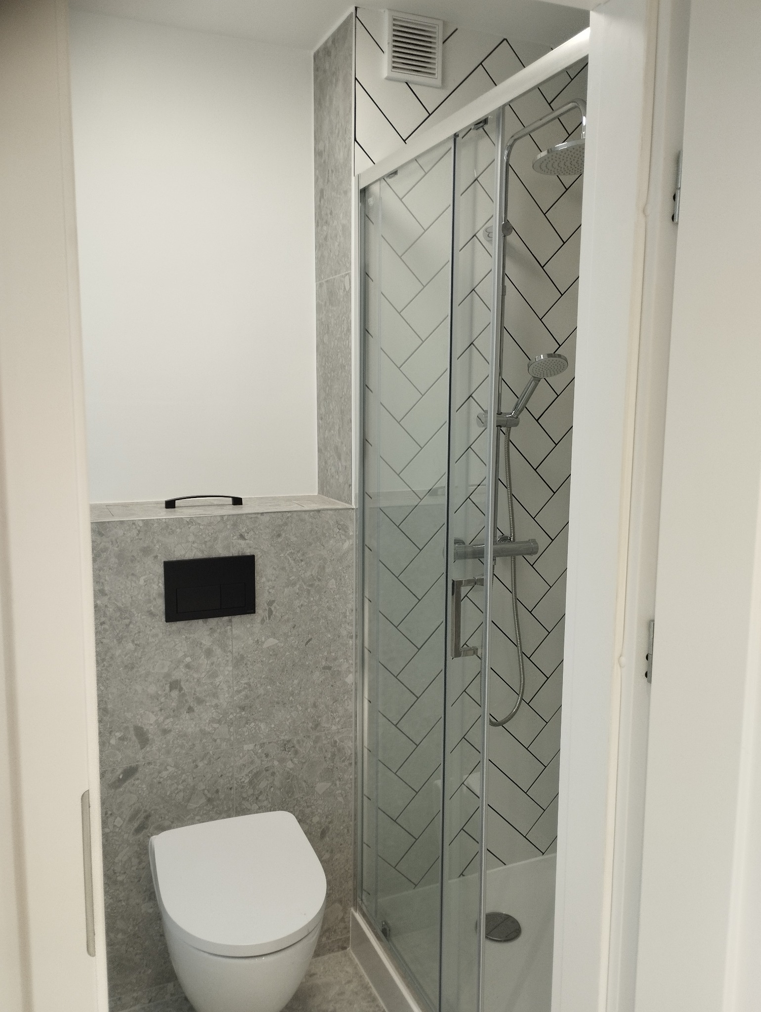 Nowoczesna łazienka po remoncie: biała toaleta, szare płytki imitujące kamień, kabina prysznicowa z wzorem w jodełkę, minimalistyczny styl, widok zza otwartych drzwi.