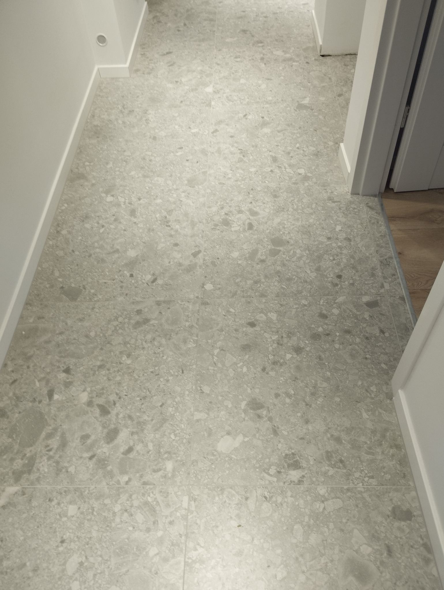 Jasnoszara, wzorzysta podłoga z płytek w korytarzu, widok z góry. Minimalistyczne białe listwy przypodłogowe i fragment ościeżnicy drzwiowej po prawej stronie.