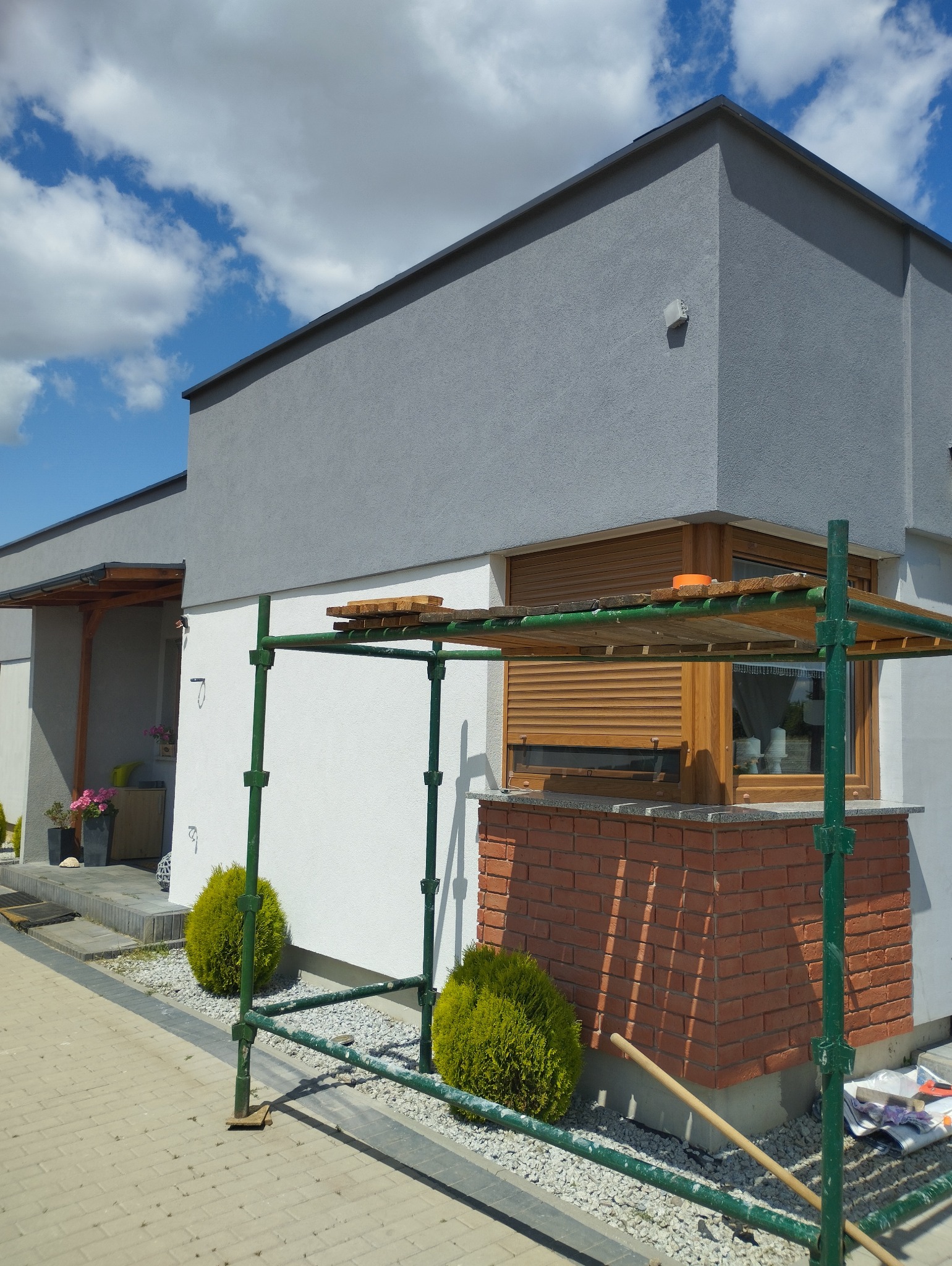 Elewacja domu w trakcie malowania: szare górne partie, białe dolne, rusztowanie, okno z drewnianymi roletami, ceglana podmurówka. Słoneczny dzień, błękitne niebo z chmurami.