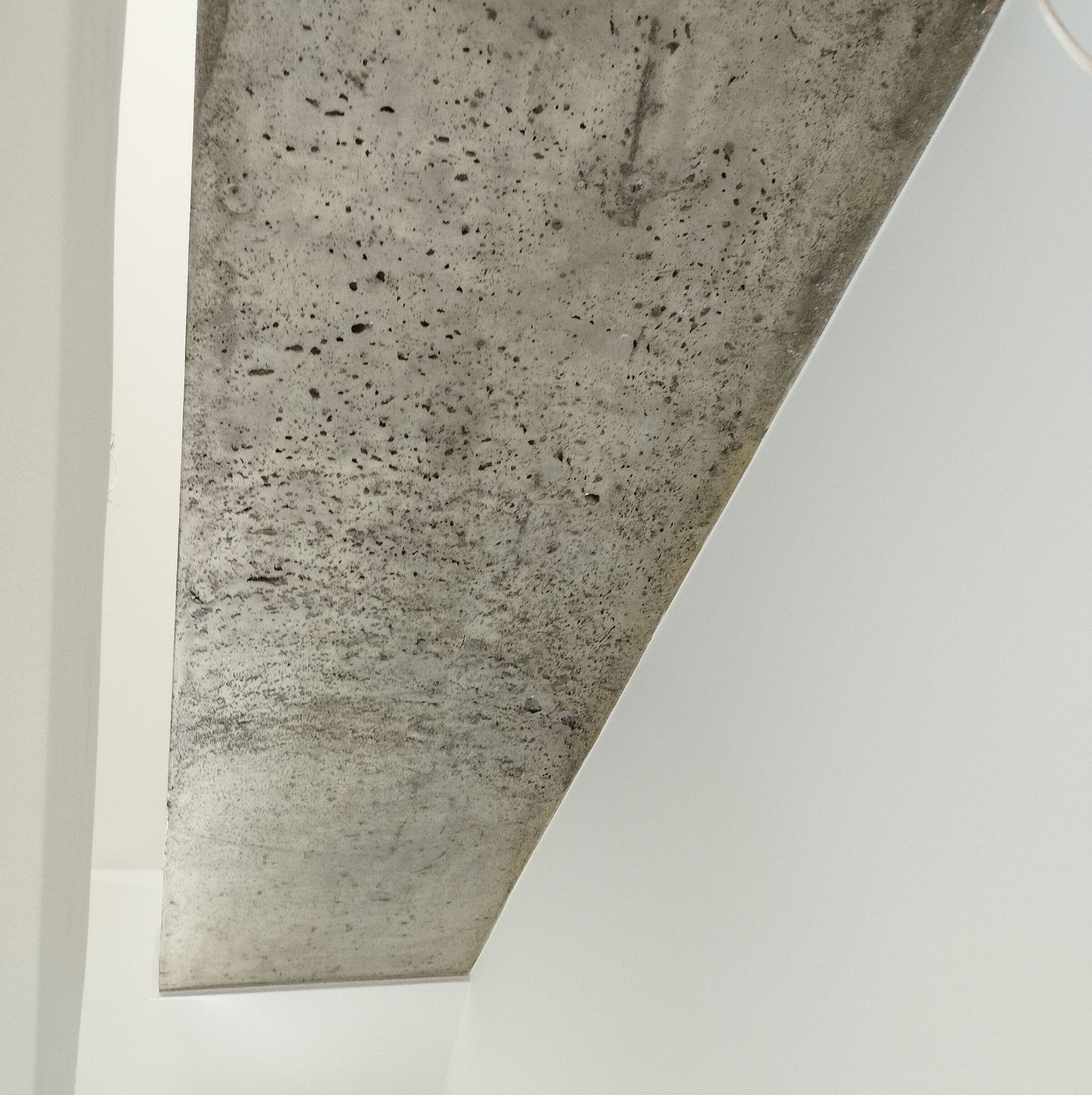 Surowy betonowy element architektoniczny kontrastuje z gładką, białą ścianą. Widoczne pory i faktura betonu, minimalistyczne ujęcie detalu wykończenia wnętrza.