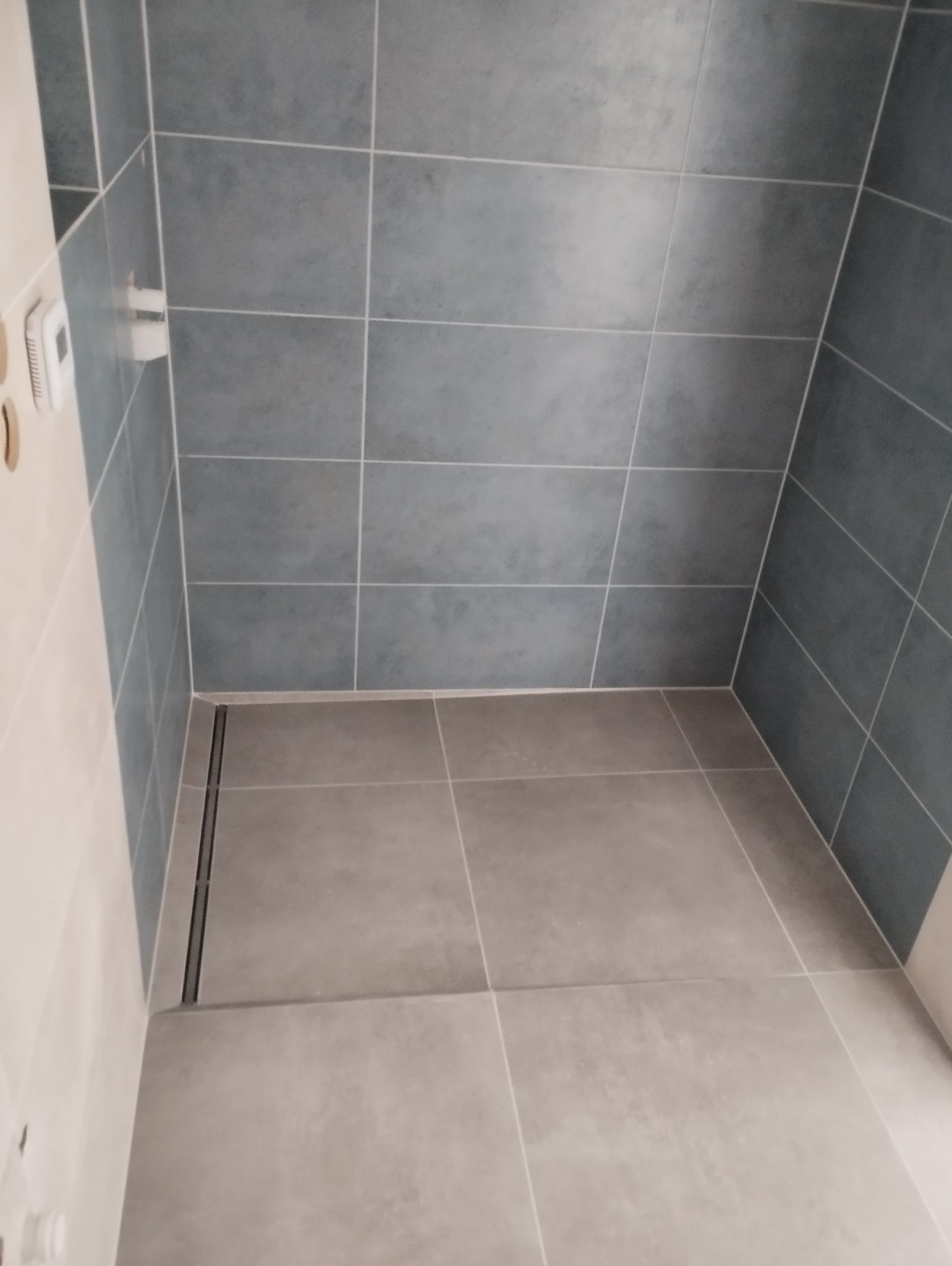 Wykończona kabina prysznicowa z szarymi płytkami na ścianach i podłodze, z odpływem liniowym. Minimalistyczny design, jasne fugi, neutralna kolorystyka.