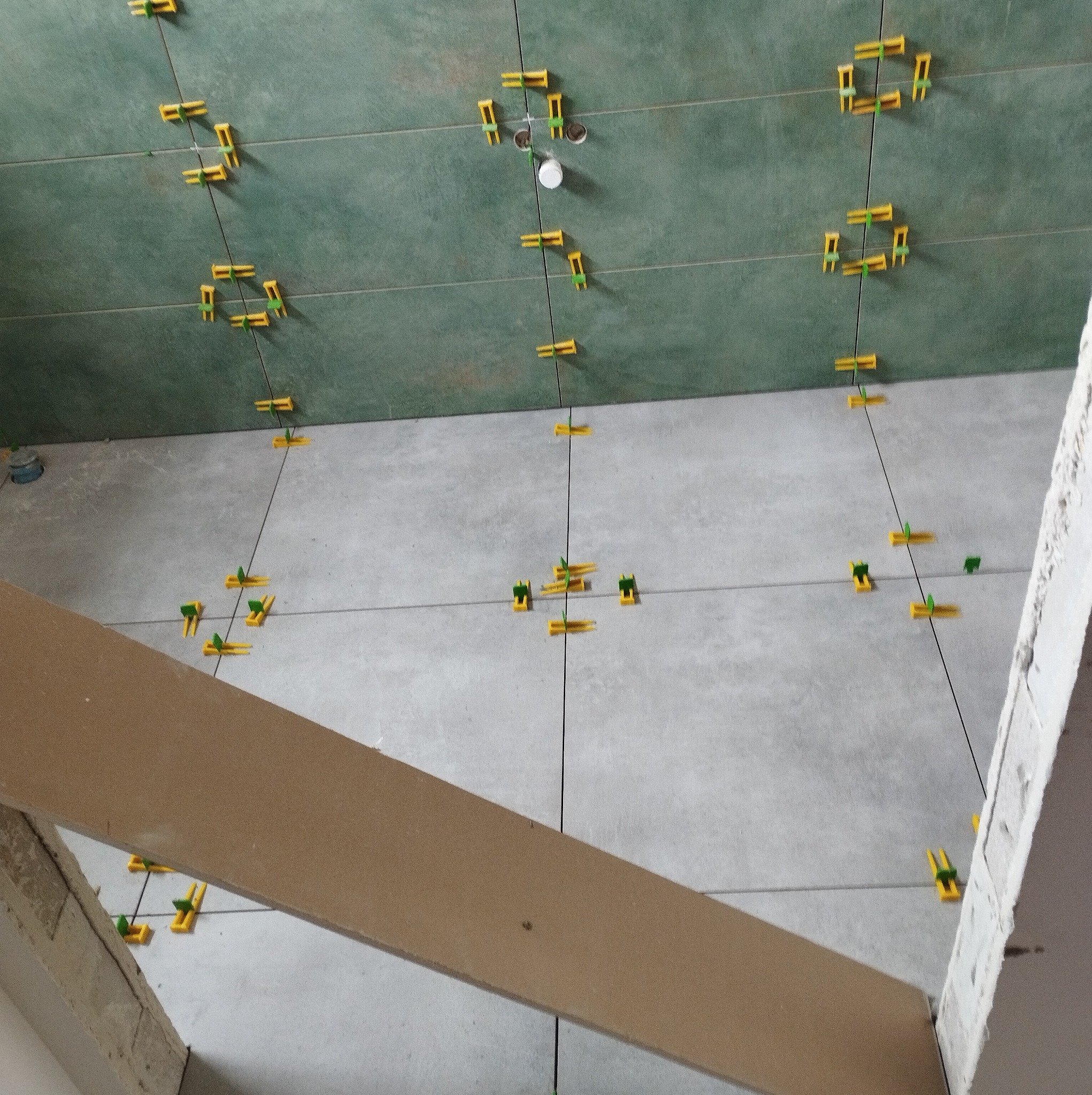 Układanie zielono-szarych płytek z żółtymi krzyżykami dystansowymi na podłodze i ścianie. Widok z góry, fragment płyty karton-gips na pierwszym planie.