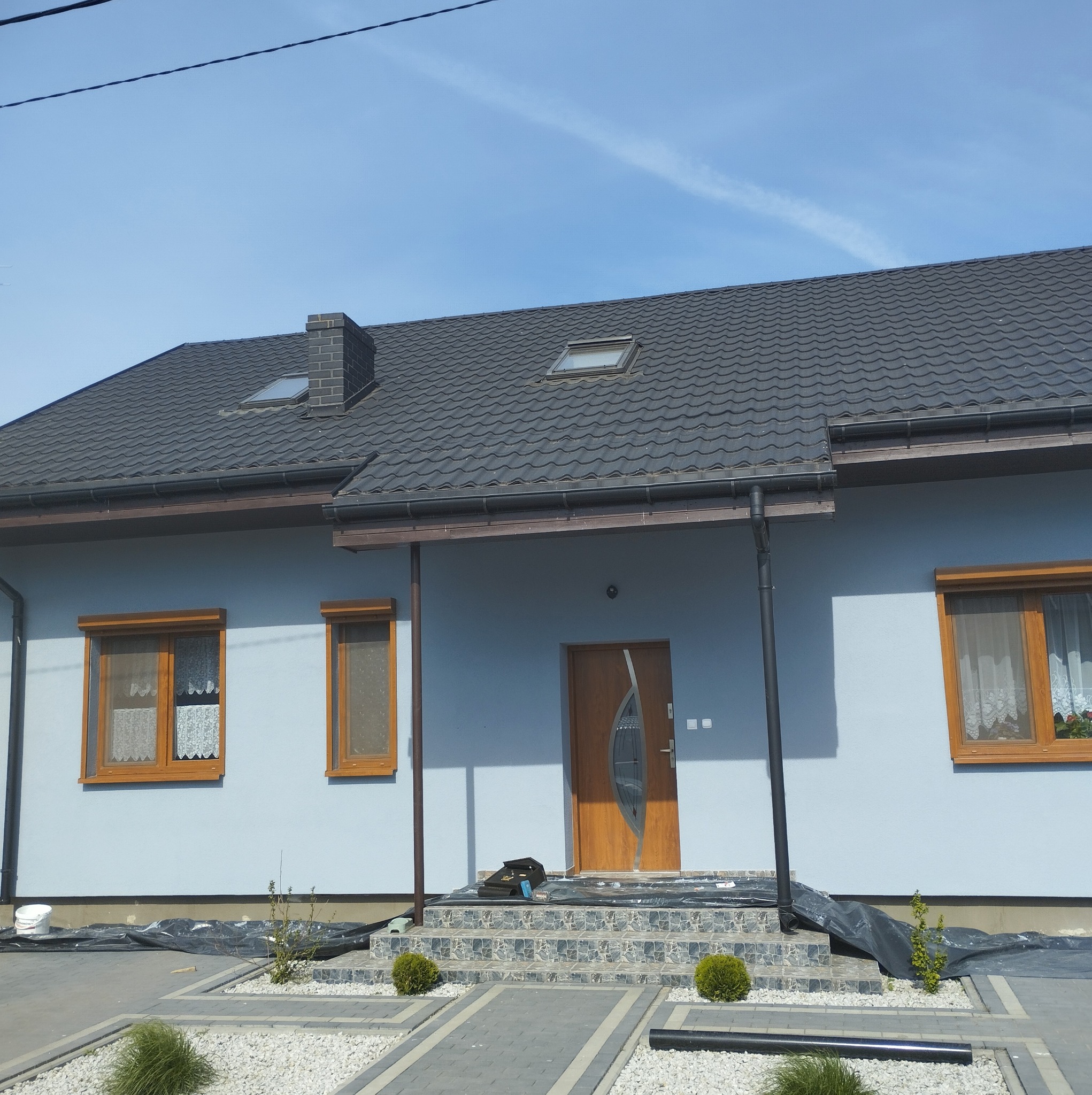 Dom jednorodzinny z szarą elewacją, brązowymi oknami i drzwiami. Widoczny dach z dachówki, okna dachowe i komin. Ułożona kostka brukowa i kamień ozdobny.