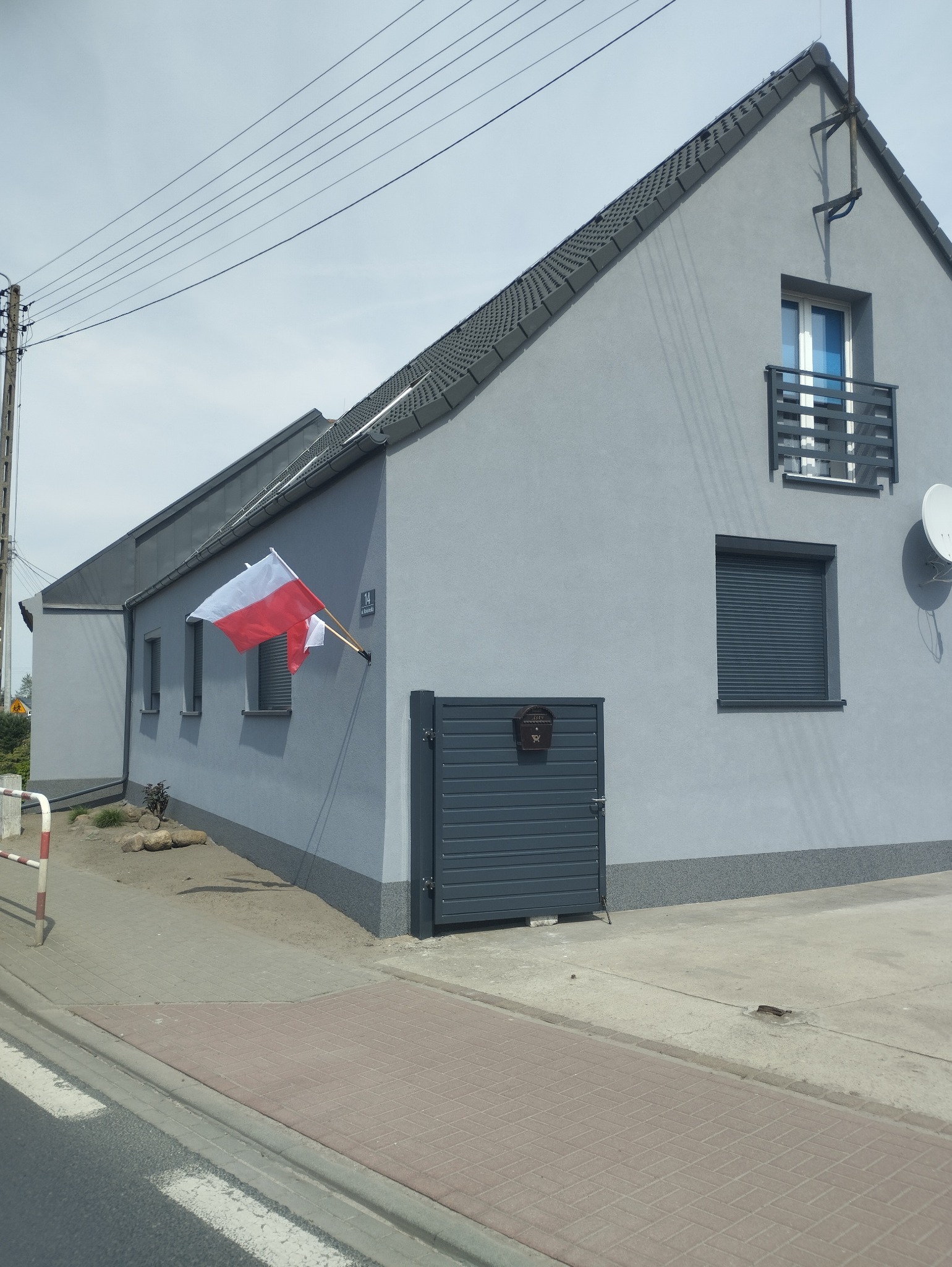 Szary dom jednorodzinny z balkonem i szarą bramą, ozdobiony polską flagą. Widoczne okna z roletami i satelita. Ujęcie z poziomu ulicy.