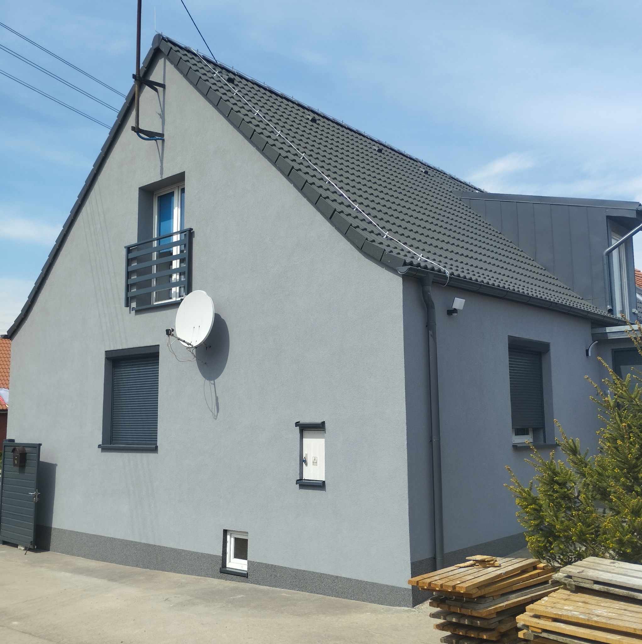 Elewacja domu jednorodzinnego w odcieniach szarości z ciemną dachówką, widoczna antena satelitarna i okna z roletami zewnętrznymi. Ujęcie z dołu, błękitne niebo.