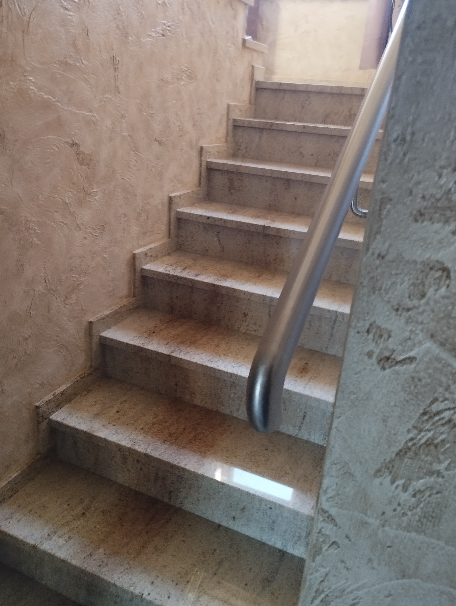 Wykończone schody wewnętrzne z kamienia w odcieniach beżu, z metalową poręczą. Ściana z dekoracyjnym tynkiem strukturalnym. Widok z dołu, oświetlenie górne.