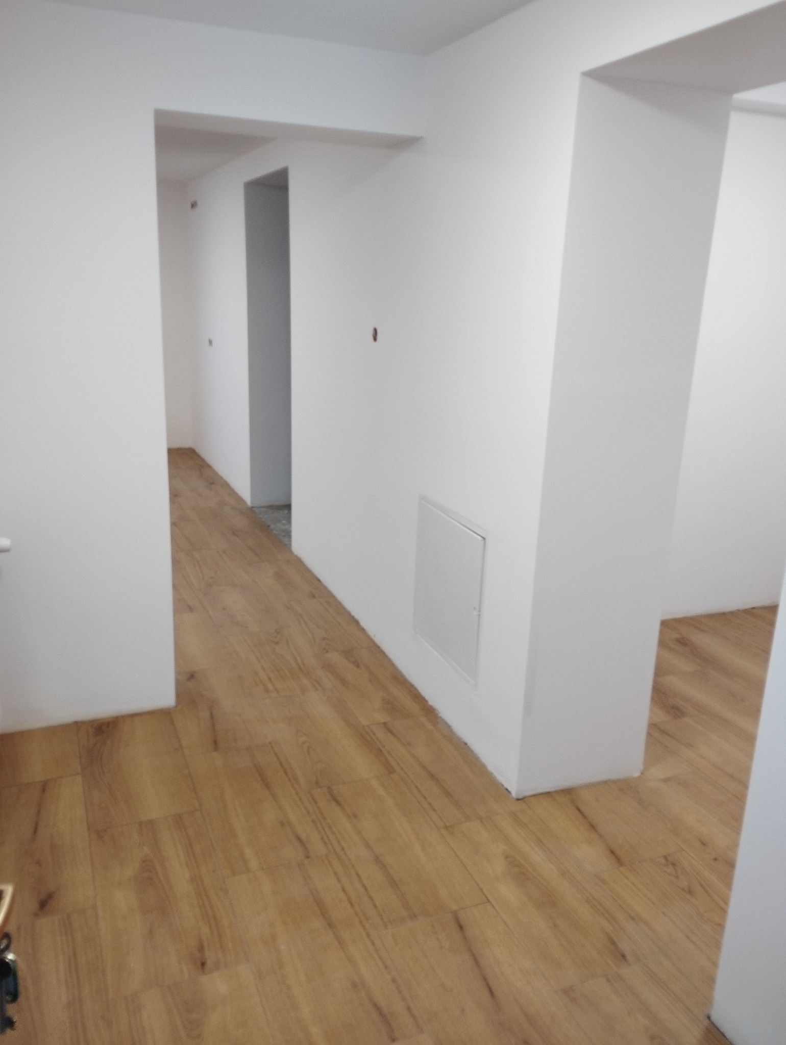 Jasne panele podłogowe w korytarzu z białymi ścianami i framugami drzwi. Widoczne przejścia do innych pomieszczeń. Minimalistyczny design, świeżo po remoncie.