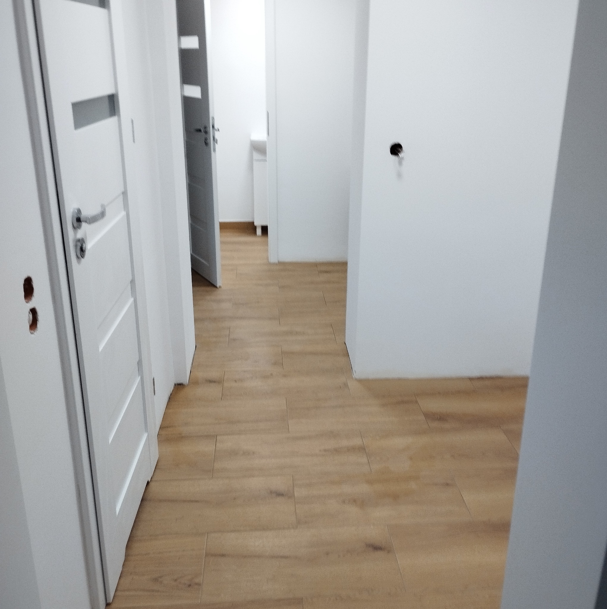 Jasne panele podłogowe w korytarzu z białymi ścianami i drzwiami. Widoczne otwory po klamkach. Otwarta przestrzeń z perspektywą na dalsze pomieszczenia.